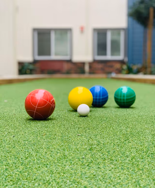 Close-up view of a bocce ball court with four bocce balls in red, yellow, blue, and green colors lined up near a small white pallino ball on green artificial turf, with a building and windows in the background.