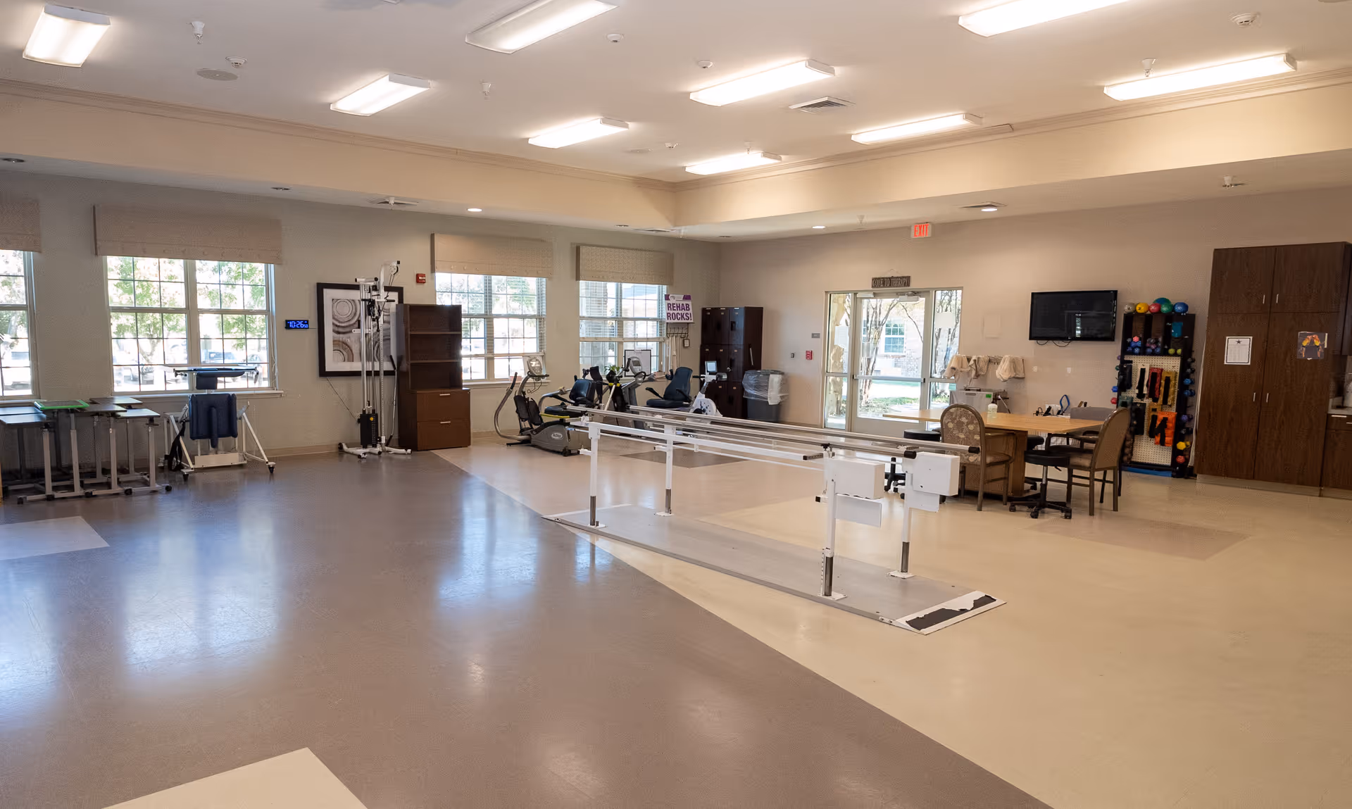 A spacious rehabilitation room with parallel bars for walking exercises in the center. The room has large windows letting in natural light, exercise equipment like stationary bikes, a table with chairs, and storage cabinets along the walls. The floor has a mix of beige and brown tiles, and the ceiling has fluorescent lighting.