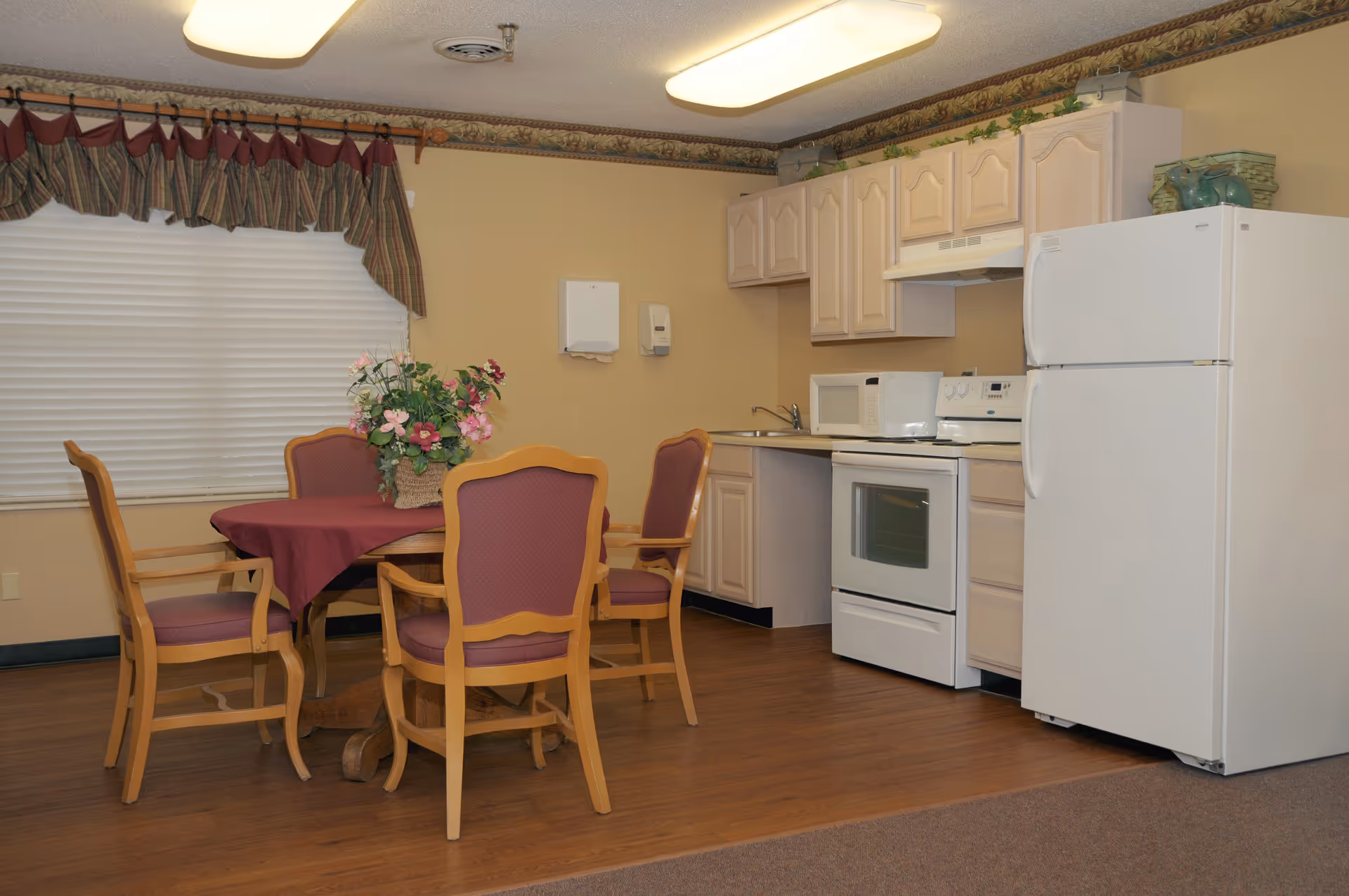 A small kitchen and dining area with a wooden table covered by a maroon tablecloth and four wooden chairs with maroon cushions. The kitchen has light-colored cabinets, a white refrigerator, a white stove with oven, a microwave, and a sink. There is a window with a valance curtain above the dining table and a floral centerpiece on the table.