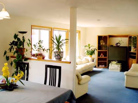 Bright living area with a dining table, white sofas, and numerous plants by the windows.