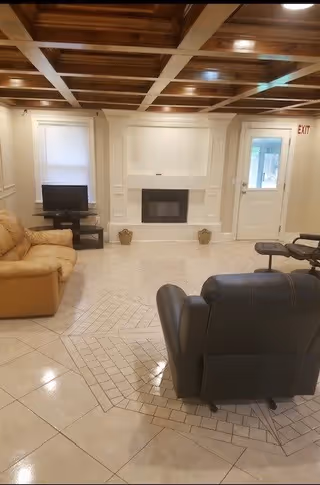 A cozy living room with a beige recliner on the left and a black leather recliner on the right. The room features a white fireplace centered on the far wall, flanked by two small decorative baskets. There is a small TV on a stand to the left of the fireplace, and a door with a window to the right. The ceiling has a wooden coffered design, and the floor is tiled with a decorative pattern in the center.