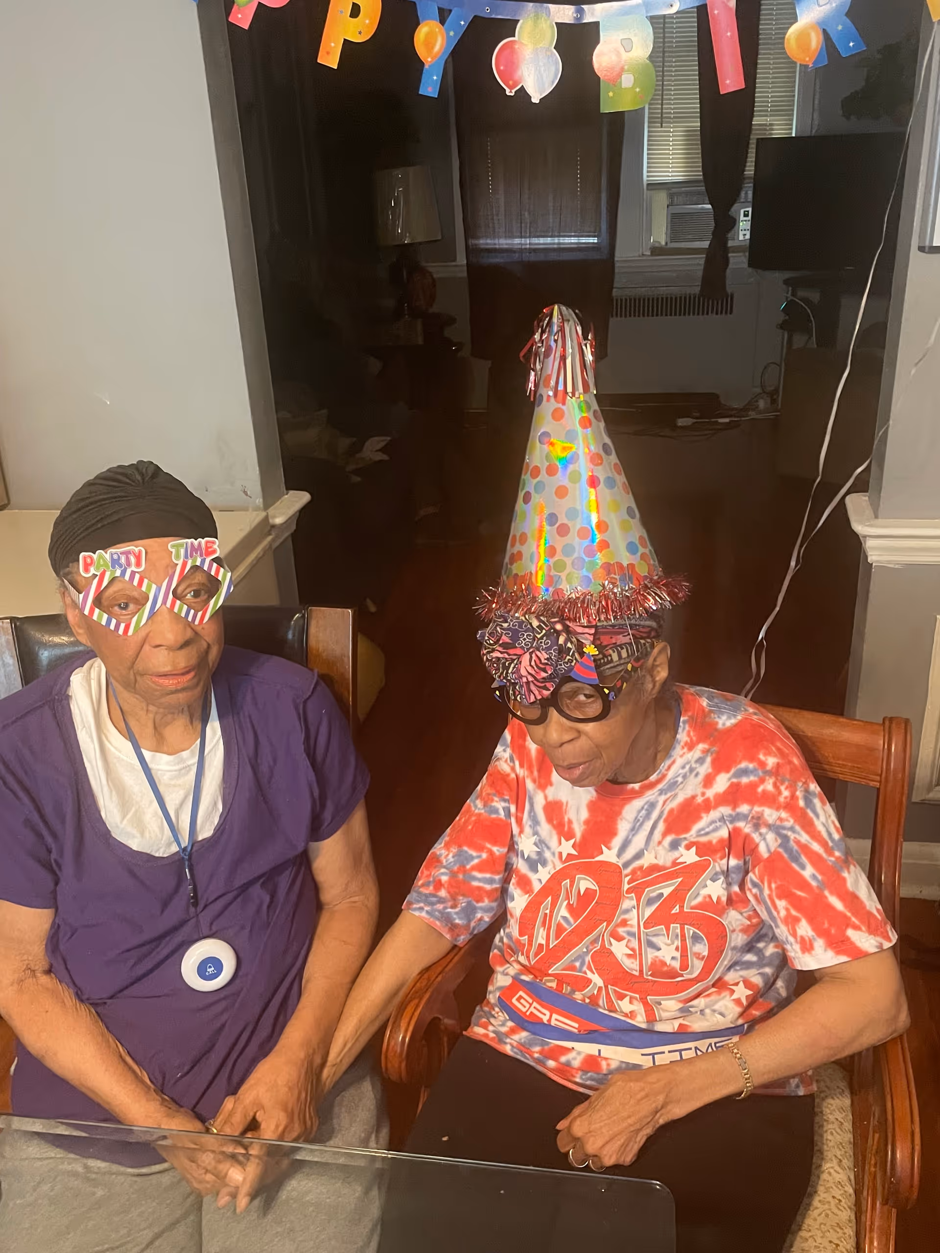 Two elderly women sitting at a table indoors, wearing festive party accessories including colorful glasses and a polka dot party hat. One woman is wearing a purple shirt and a black head wrap, while the other is wearing a red, white, and blue tie-dye shirt and a patterned head wrap. A 'Happy Birthday' banner is partially visible hanging above them.