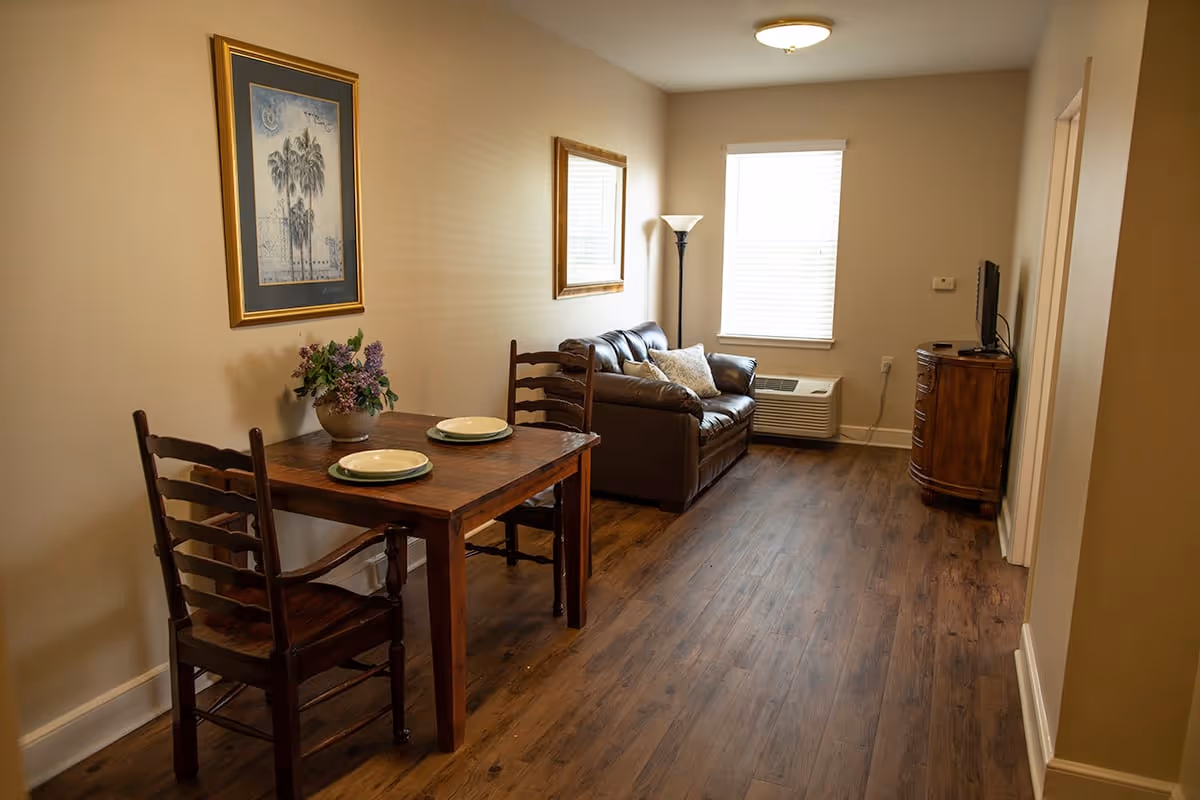 A small living area with a wooden table set for two with plates and a vase of flowers, a brown leather loveseat with two pillows, a floor lamp, a window with blinds, and a wooden cabinet with a TV on top. The room has wooden flooring and beige walls with framed artwork and a mirror.