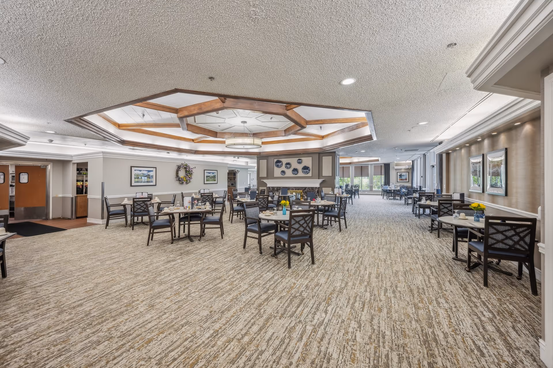 Spacious dining room with multiple tables and chairs arranged neatly. The room features a decorative ceiling with wooden beams and a central light fixture. There are framed pictures and a wreath on the walls, and large windows letting in natural light at the far end. The carpeted floor and overall decor create a warm and inviting atmosphere.