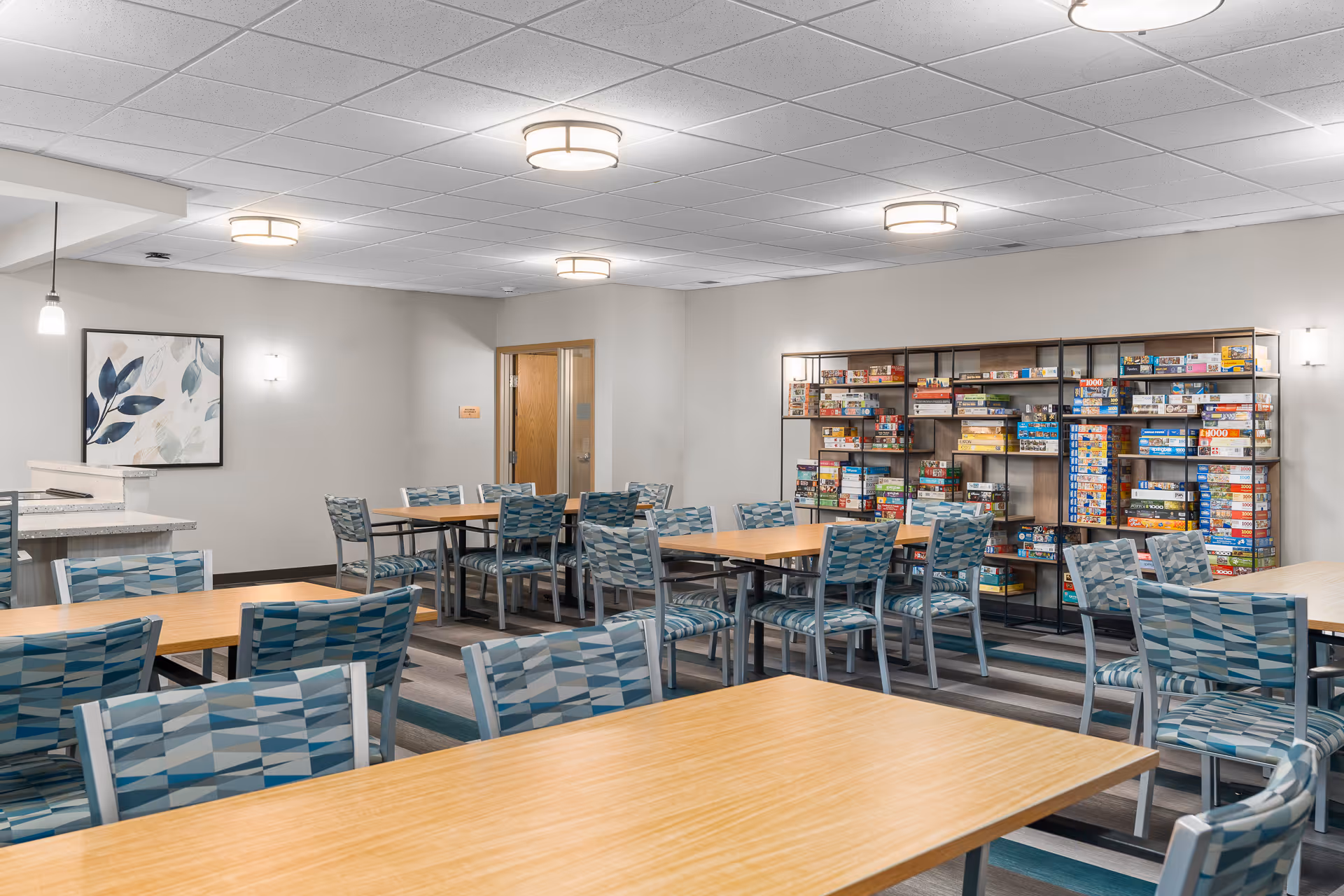 A well-lit community room with multiple wooden tables and patterned chairs arranged for group activities. Along one wall, there is a large shelving unit filled with various board games. The room has a neutral color scheme with light gray walls, a framed abstract leaf artwork, and modern ceiling lights.