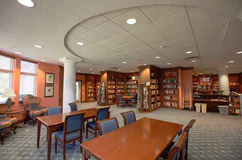 A spacious library room with wooden bookshelves filled with books along the walls. Several wooden tables with blue cushioned chairs are arranged in the center of the room. The room has large windows with blinds allowing natural light to enter, and there are two white columns supporting the ceiling. A comfortable armchair and a lamp are positioned near the windows.