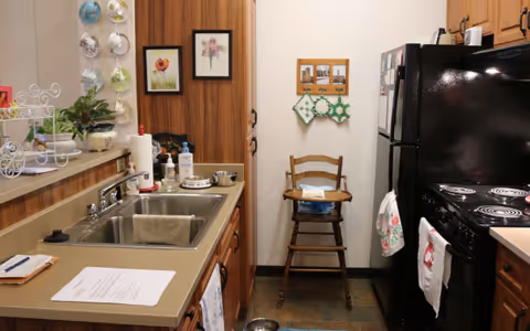Compact kitchen with a double sink, countertop items, wooden cabinets, black refrigerator and stove, and a wooden high chair against a white wall with framed art.