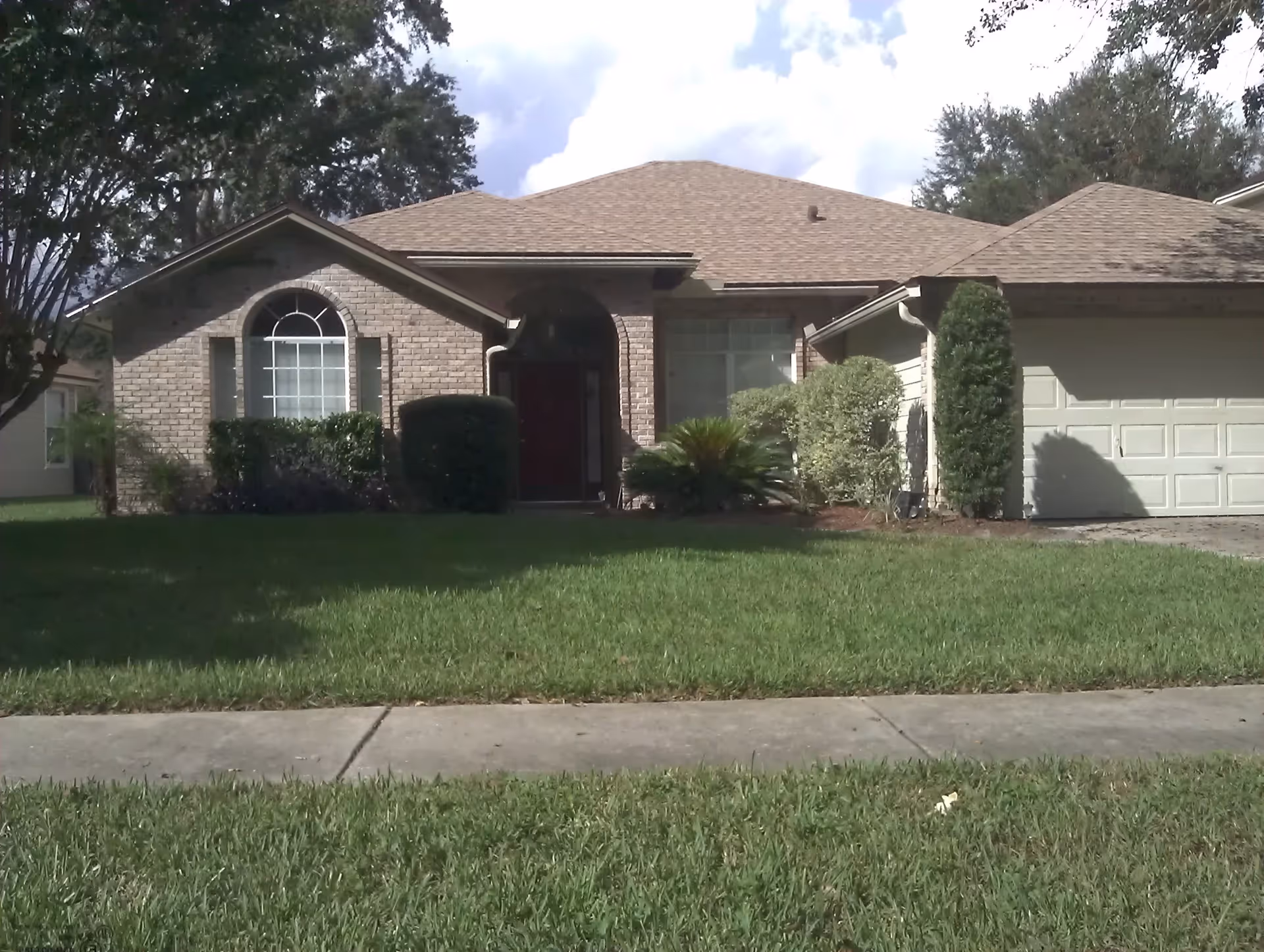 Front exterior view of a single-story brick house with a well-maintained lawn, bushes, and trees under a partly cloudy sky.