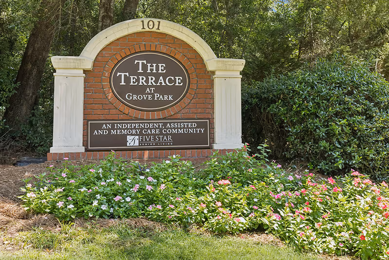 A brick and stone sign for The Terrace at Grove Park, an independent, assisted and memory care community by Five Star Senior Living, surrounded by green bushes and pink flowers with trees in the background.