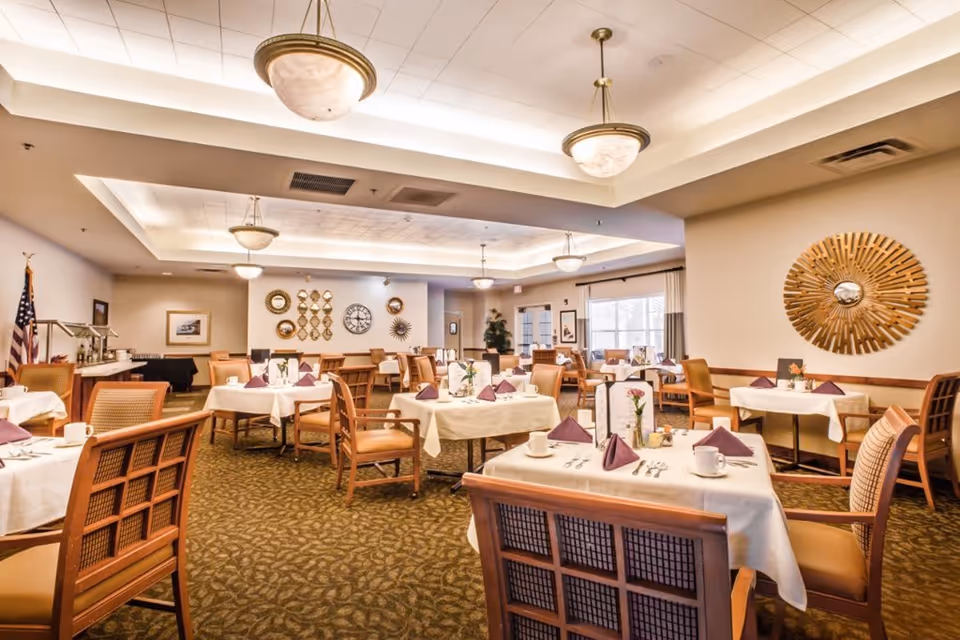 A spacious dining room with multiple tables covered in white tablecloths, each set with purple folded napkins, cups, and cutlery. The room features warm lighting from ceiling fixtures, decorative wall mirrors, framed pictures, and an American flag in the corner. The carpet has a leafy pattern, and large windows allow natural light to enter.