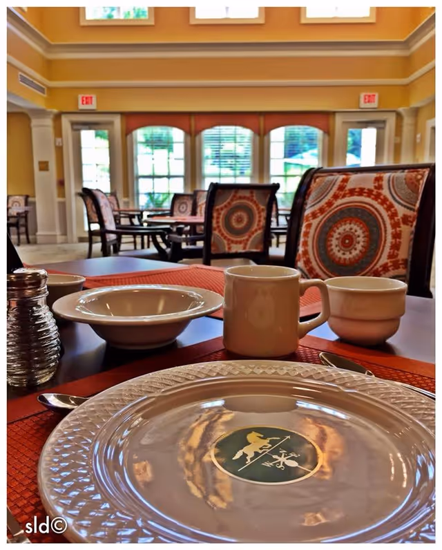 A dining table set with a white plate featuring a logo, a bowl, a coffee mug, a small cup, a spoon, and a salt shaker in a senior living facility dining room. The background shows chairs with patterned upholstery and large windows letting in natural light.