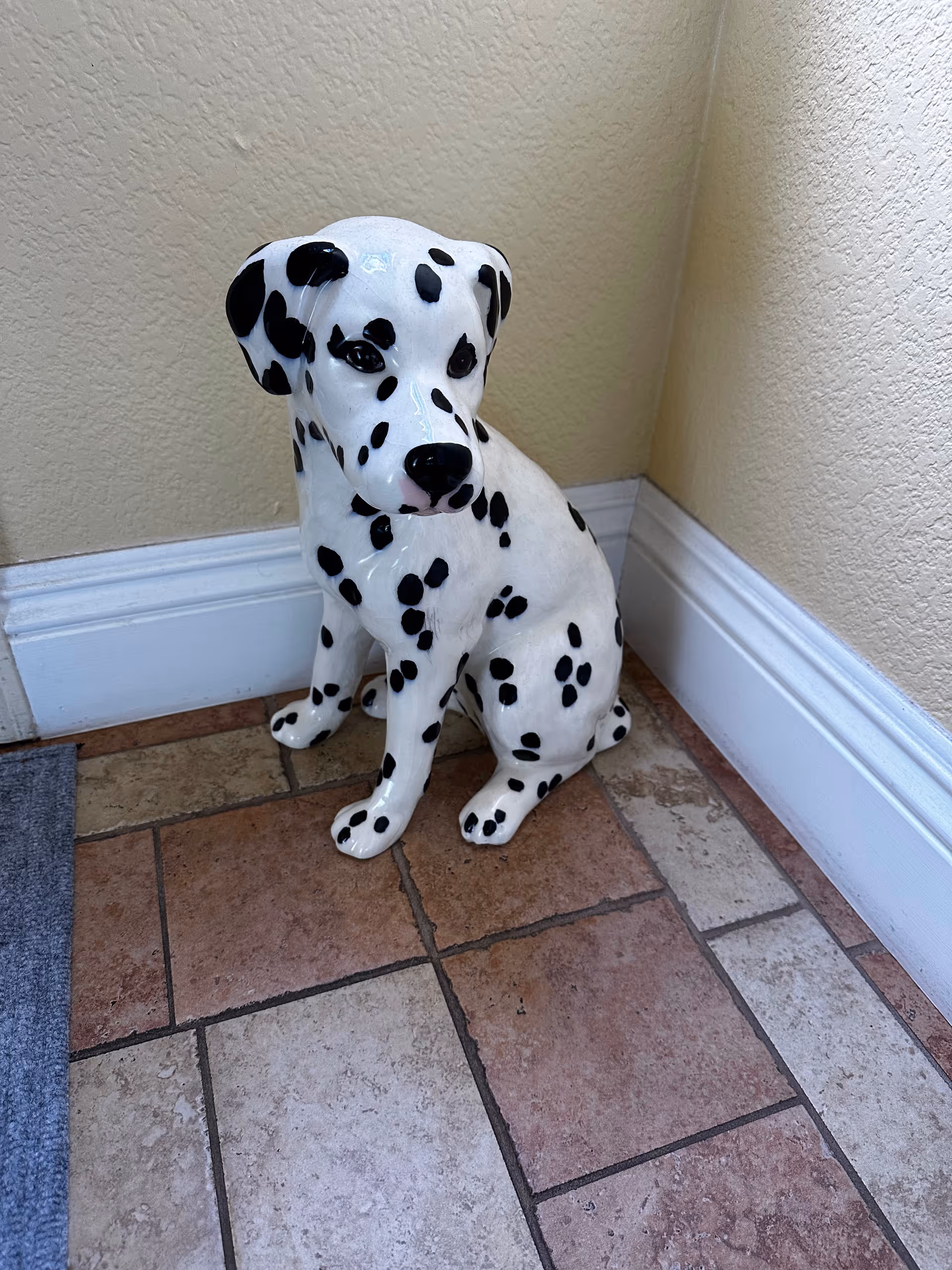 A ceramic statue of a Dalmatian dog with black spots sitting on a tiled floor in the corner of a room with beige textured walls and white baseboards.
