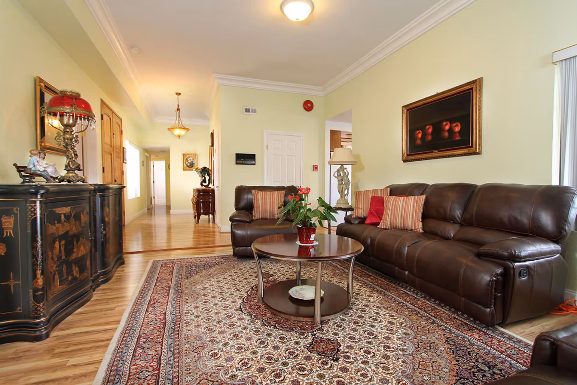 Bright living room with a brown leather sofa, round glass-top coffee table on a patterned rug, and decorative furniture along a hallway.