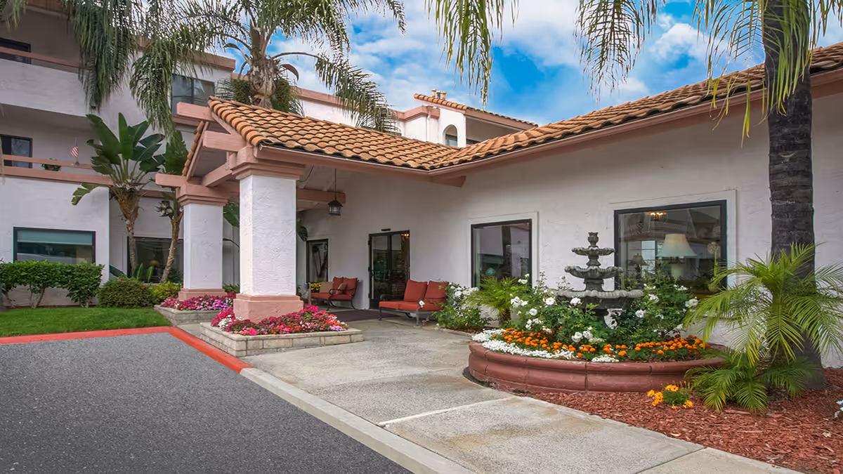 Covered entrance of a residential building with a tiled roof, outdoor seating, a tiered fountain and colorful flower beds.