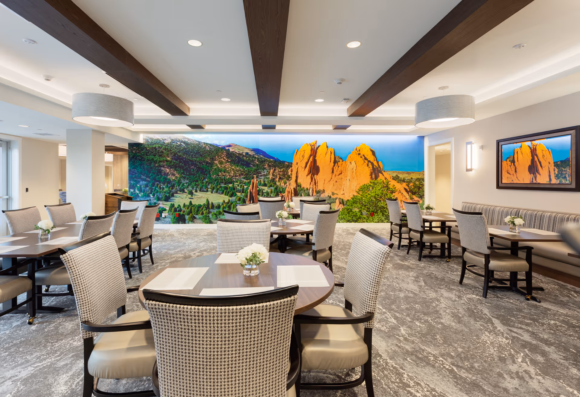 A spacious dining room with multiple round and rectangular tables surrounded by cushioned chairs. The room features a large, vibrant mural of a mountainous landscape on one wall and a matching framed picture on another wall. The ceiling has wooden beams and modern light fixtures, and the floor is covered with a patterned carpet.