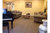 A cozy living room area with two striped sofas and one patterned sofa arranged around a wooden coffee table. A black piano is visible on the left side, and a large decorative vase is on the right. The walls are painted light yellow, and a framed picture hangs on the wall.