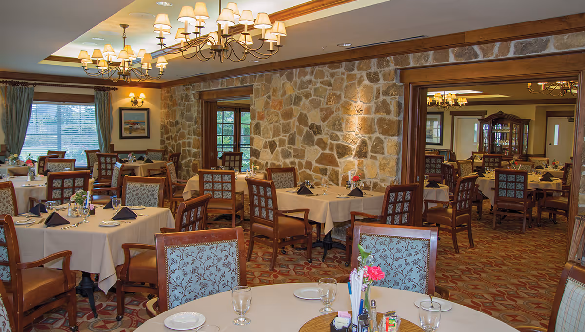 A spacious dining room with multiple tables covered in beige tablecloths, each set with plates, glasses, and black napkins. The chairs have wooden frames with patterned upholstery. The room features stone accent walls, large windows with curtains, chandeliers with multiple lampshades, and a carpeted floor with a geometric pattern. There are floral centerpieces on the tables and a wooden cabinet with glass doors in the background.