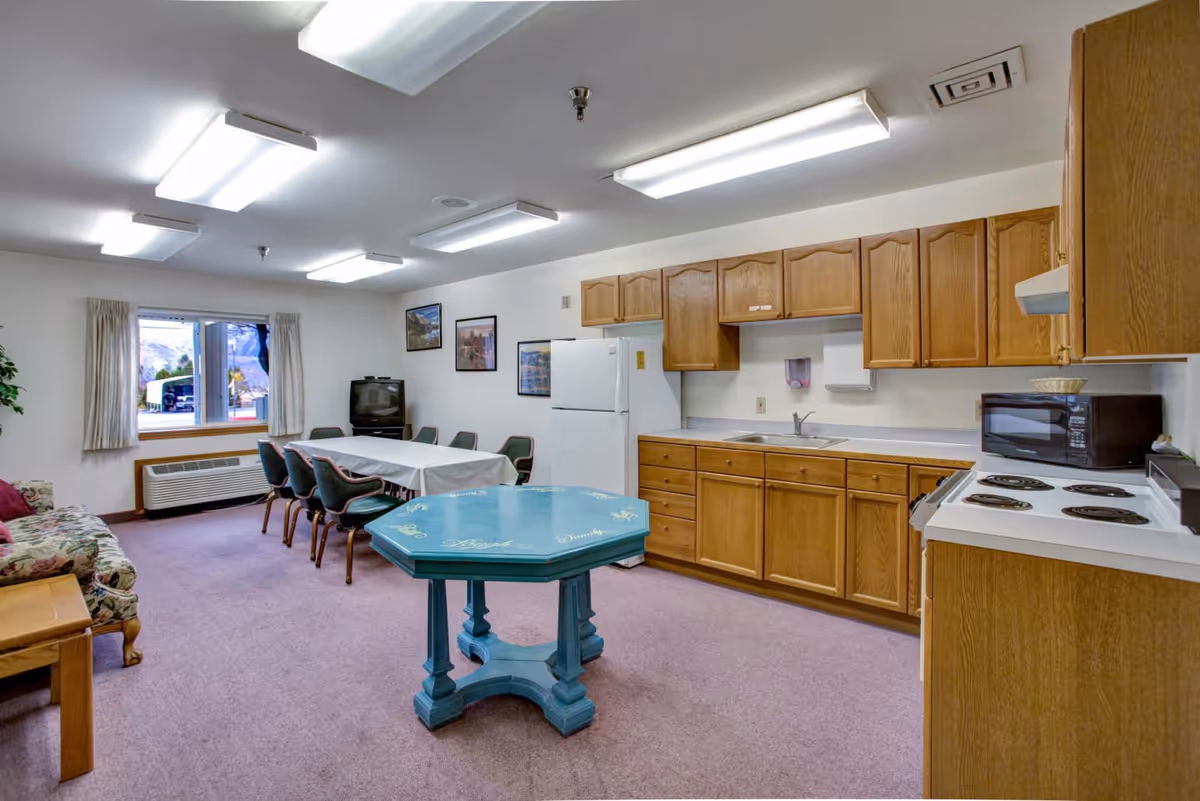 Interior view of a senior living facility common room featuring a kitchen area with wooden cabinets, a white refrigerator, microwave, and stove. There is a teal octagonal table in the center of the room, a dining table with six chairs covered with a white tablecloth near a window, and a floral-patterned sofa with a wooden side table. The room is carpeted and well-lit with ceiling fluorescent lights.
