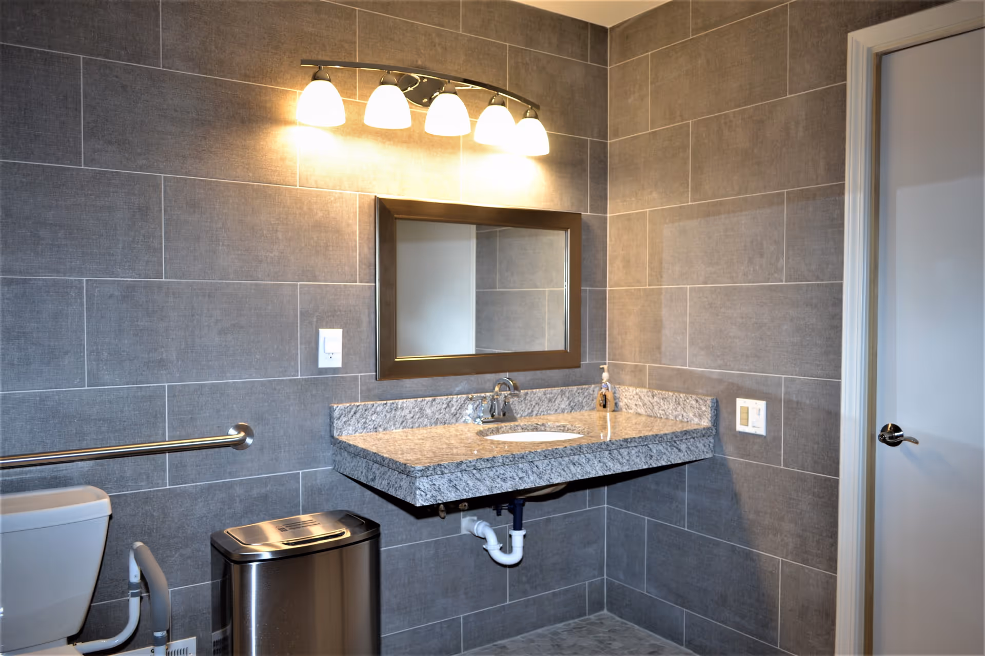 A modern bathroom with gray tiled walls, a floating granite countertop with an under-mount sink, a rectangular mirror above the sink, a four-light fixture mounted above the mirror, a stainless steel trash can, a toilet with grab bars, and a closed white door with a silver handle.