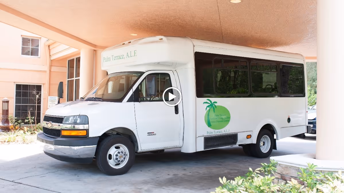 A white shuttle bus parked under a covered entrance of a building. The bus has the logo and name of Palm Terrace Assisted Living Facility & Adult Day Care Center on its side and front. The building exterior is light peach with windows and columns visible.