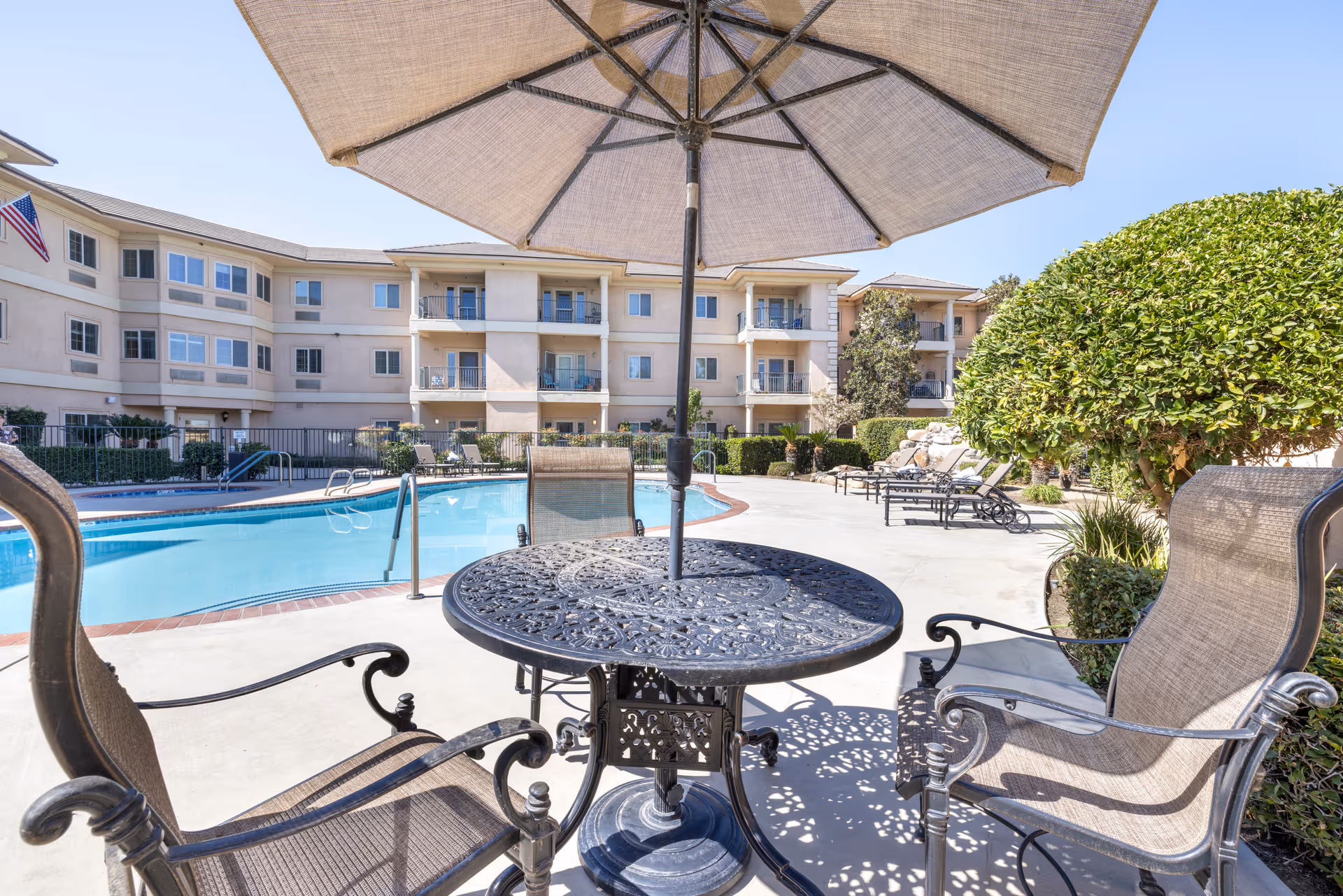 Outdoor pool area at Brookdale Riverwalk with a round metal table and three chairs under a large beige umbrella in the foreground. The pool is surrounded by lounge chairs and greenery, with a multi-story residential building in the background under a clear blue sky.