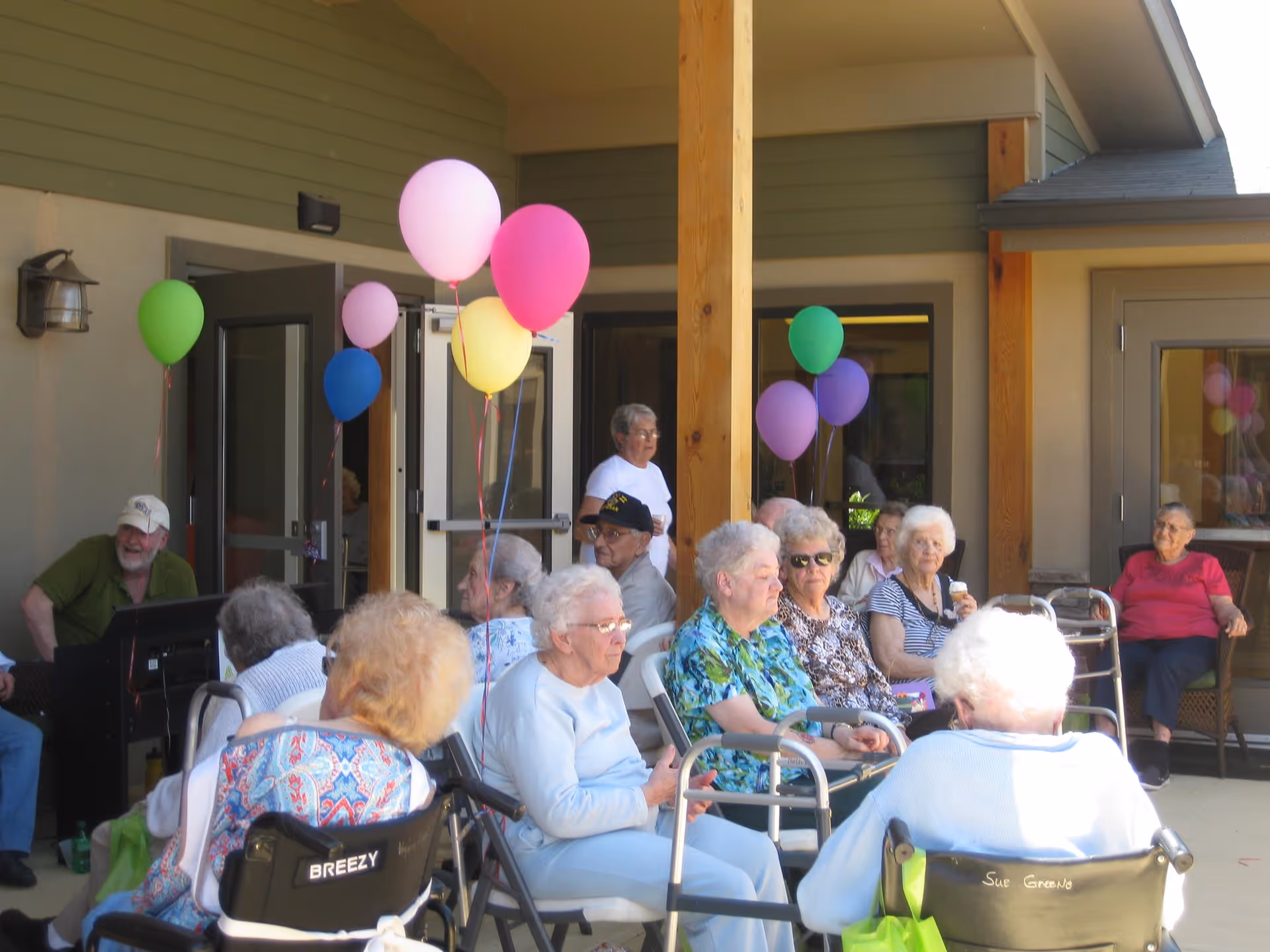 A group of elderly people sitting outside on a covered patio area, some in wheelchairs and others in chairs. Colorful balloons in pink, purple, green, yellow, and blue are tied to the chairs and posts. The setting appears to be a social gathering at a senior living facility with a man playing a keyboard in the background.