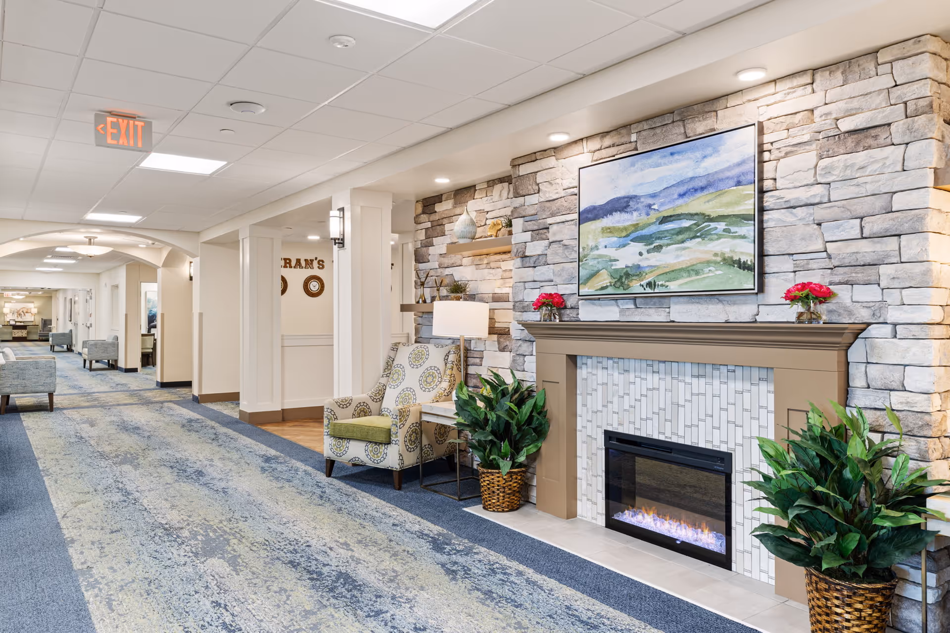 A well-lit hallway in a senior living facility featuring a stone accent wall with an electric fireplace, a framed landscape painting above it, and two potted plants on either side. There is a patterned armchair with a side table and lamp next to the fireplace. The hallway has a blue and green carpet, white walls, and ceiling lights, with an exit sign visible on the ceiling.