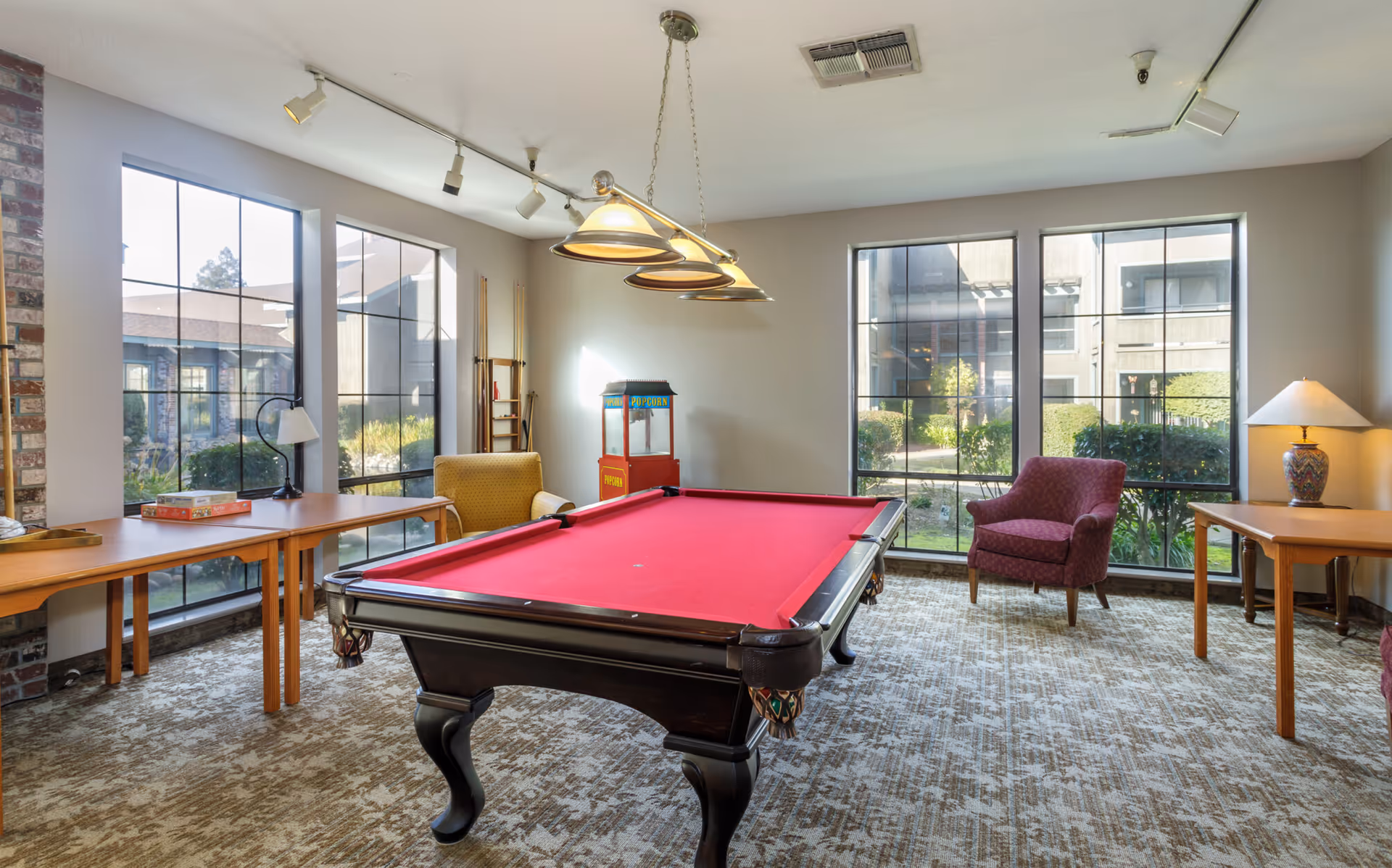 Bright recreation room with a red-felt pool table centered, large windows overlooking a courtyard, seating, and game tables.