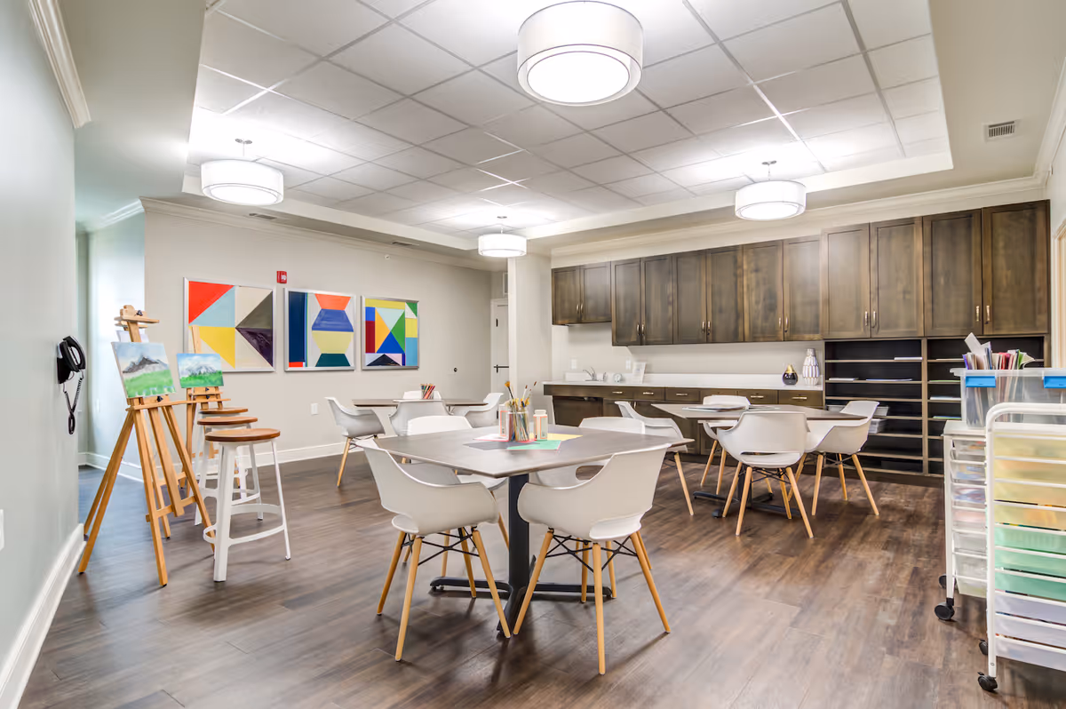 A bright and spacious art activity room with wooden floors, white modern chairs around tables, easels with paintings, colorful geometric artwork on the wall, and dark wooden cabinets along the back wall. The room is well-lit with ceiling lights and has art supplies on the tables and in storage carts.