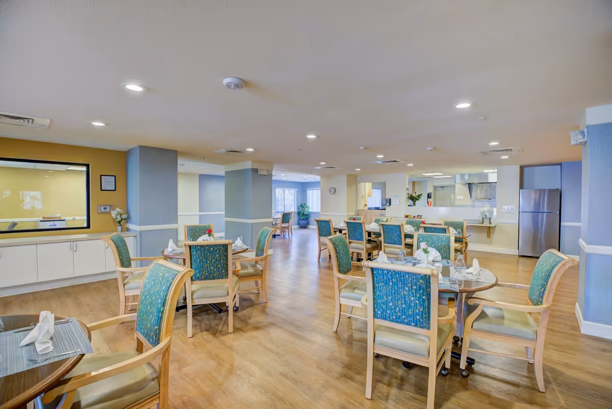 A bright and spacious dining room in a senior living facility with multiple round tables set with napkins and glassware. The room features wooden flooring, green upholstered chairs with wooden frames, and a kitchen area with a stainless steel refrigerator in the background. The walls are painted in soft blue and yellow tones, and the ceiling has recessed lighting.