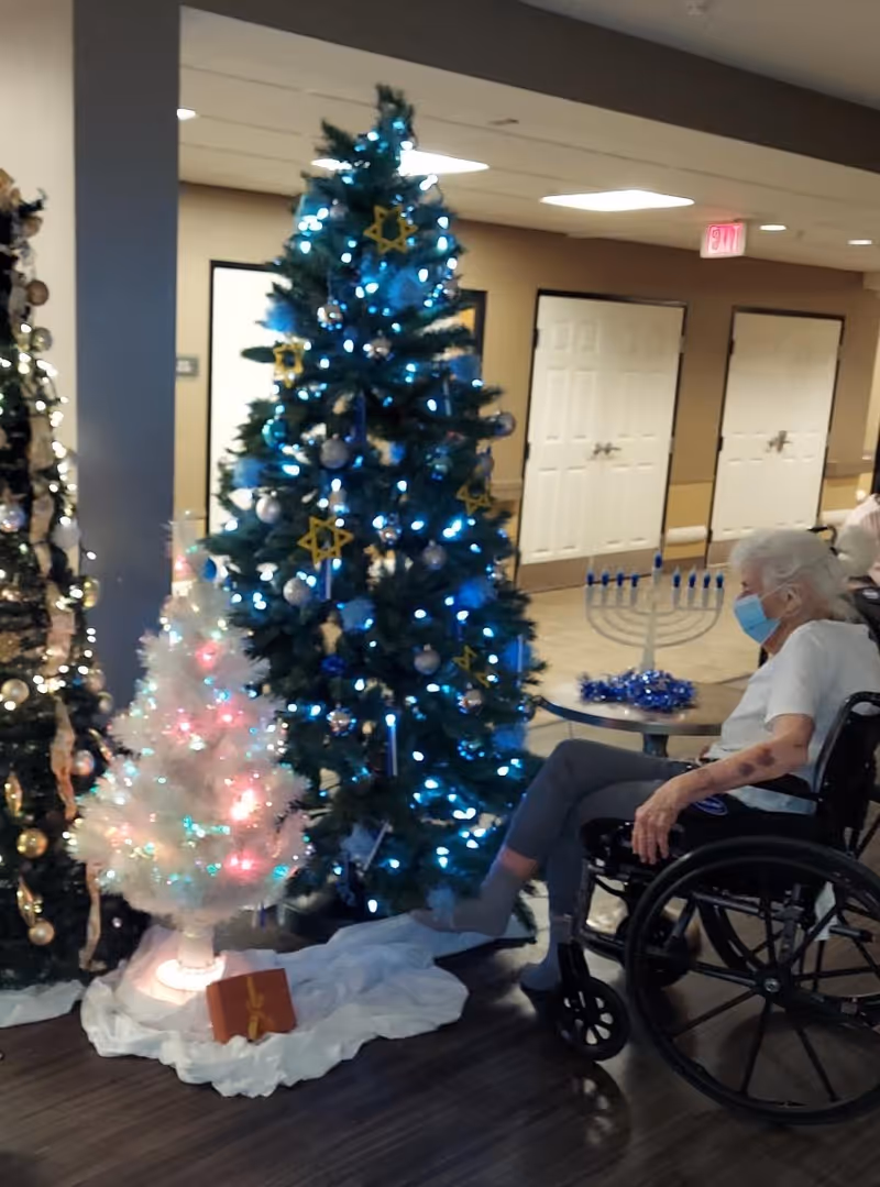 An elderly woman wearing a face mask sits in a wheelchair in a hallway decorated with multiple Christmas trees adorned with lights and ornaments. A menorah is placed on a small table nearby, indicating a celebration of both Christmas and Hanukkah. The setting appears to be inside a senior living facility with beige walls and white double doors in the background.