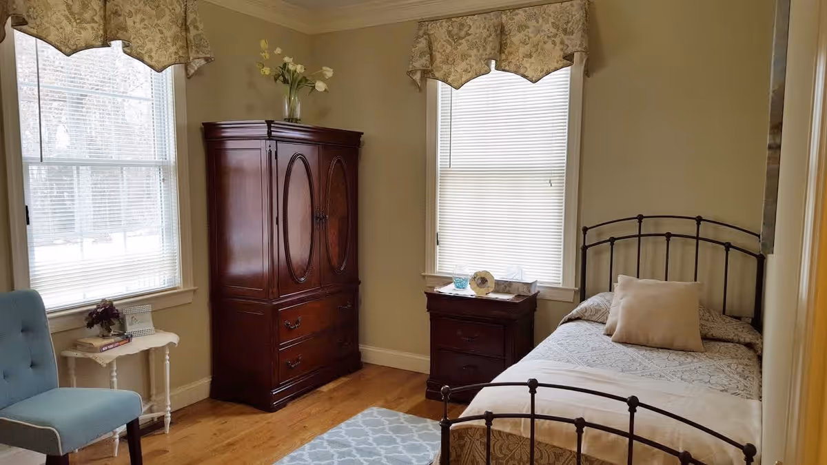 A cozy bedroom with a single bed featuring a metal frame and a beige pillow. There is a wooden nightstand with a decorative plate and a tissue box next to the bed. A large wooden armoire stands against the wall between two windows with blinds and floral valances. A small white side table with books and a flower vase is next to a blue upholstered chair. The room has wooden flooring and beige walls.