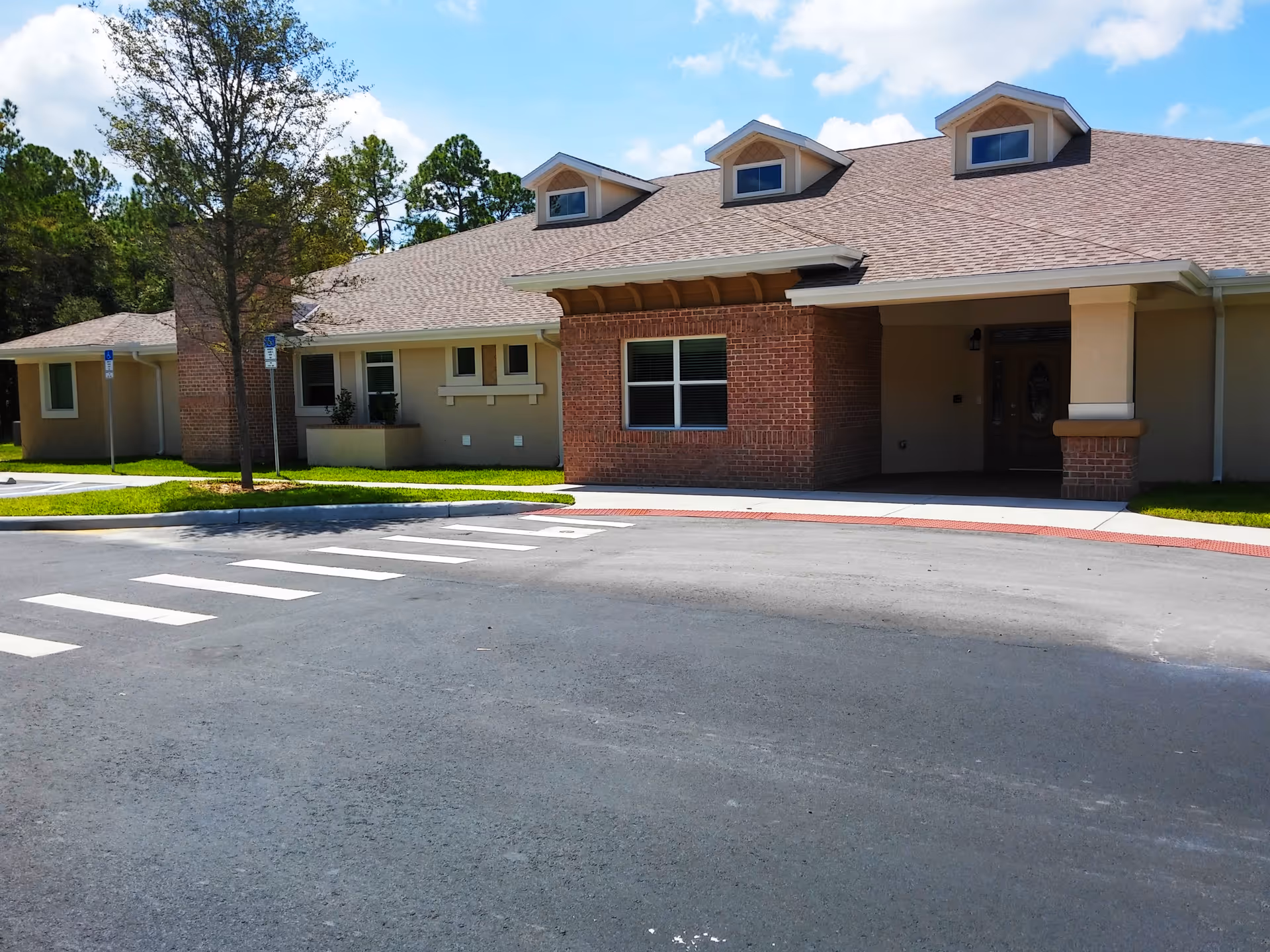 Exterior view of a single-story senior living facility building with a brick and beige facade, three dormer windows on the roof, a covered entrance, a tree, and a crosswalk in front of the building under a partly cloudy sky.