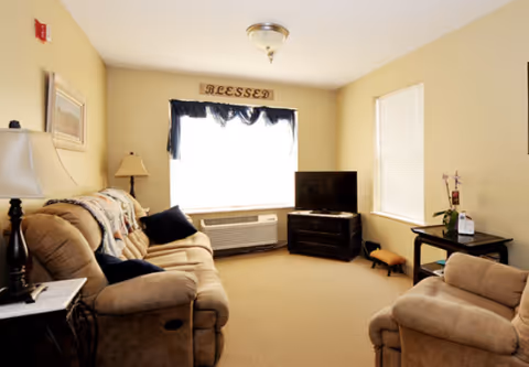 A cozy living room with beige walls and carpet, featuring a tan sofa and armchair with dark blue cushions, a small black TV on a wooden stand, a window with a dark blue valance, a side table with a lamp, and a decorative sign above the window that reads 'BLESSED'.