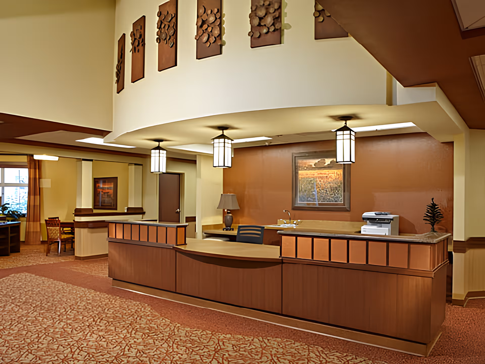 Reception desk area in a senior living facility with warm lighting, a curved wooden counter, three hanging lantern-style lights, a framed landscape painting on the wall, a lamp, a printer, and decorative wall art above the desk. The carpet has a patterned design and there is a seating area visible in the background near a window with curtains.