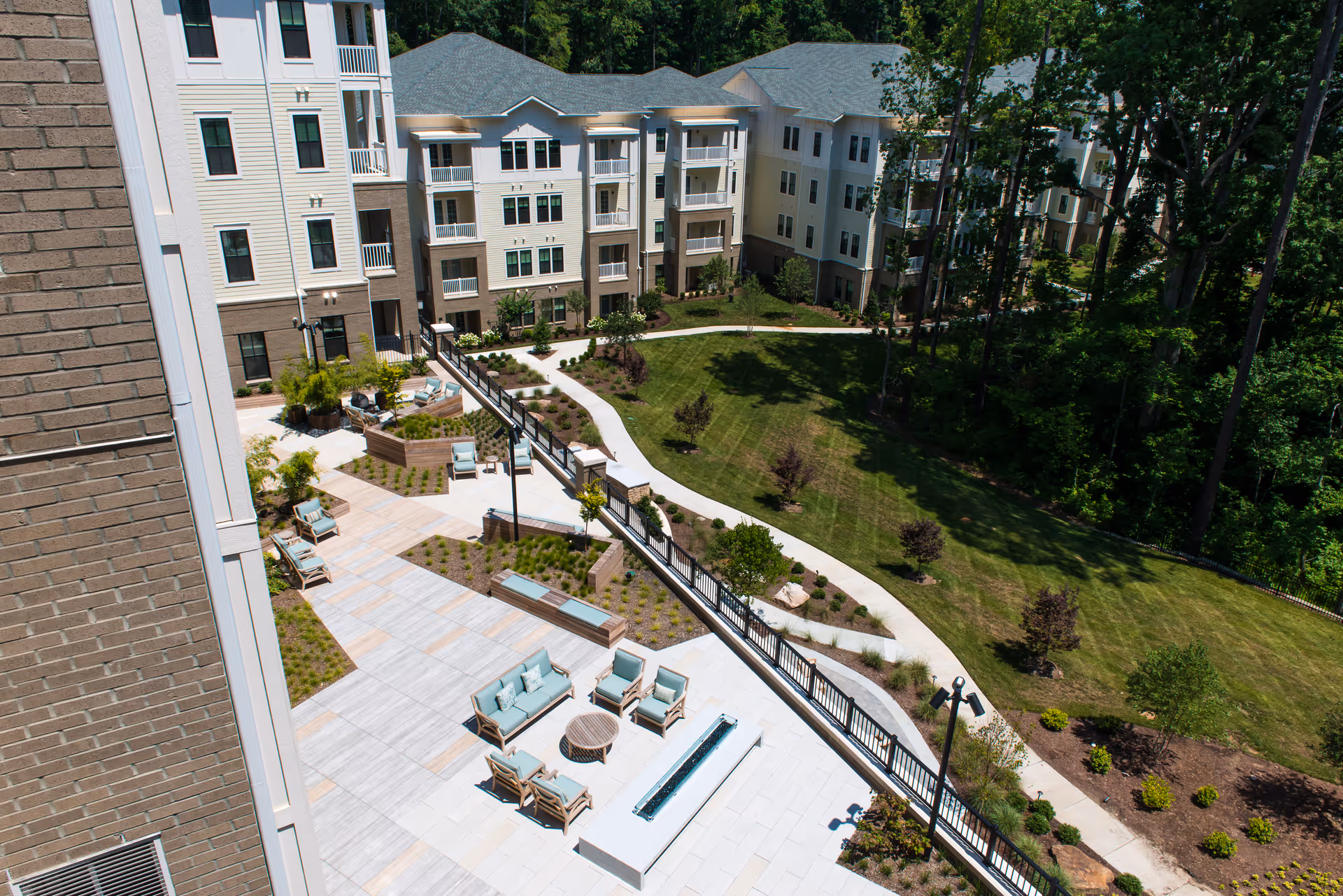 Aerial view of an outdoor patio area at The Templeton of Cary featuring seating arrangements with chairs and sofas around a fire pit, surrounded by landscaped gardens and pathways, adjacent to a multi-story residential building with balconies.