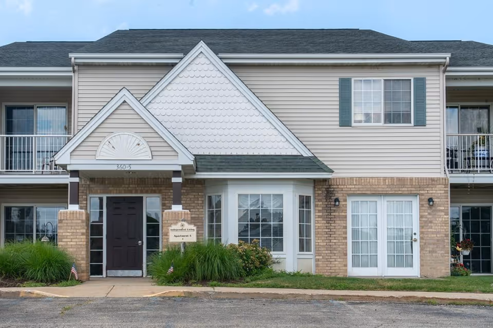 Exterior view of a two-story residential building with beige siding and brick accents. The building has a dark front door under a small gabled roof with white trim and a sign that reads 'Independent Living Apartment 5'. There are windows with white frames and green shutters, small bushes, and grass in front of the building.