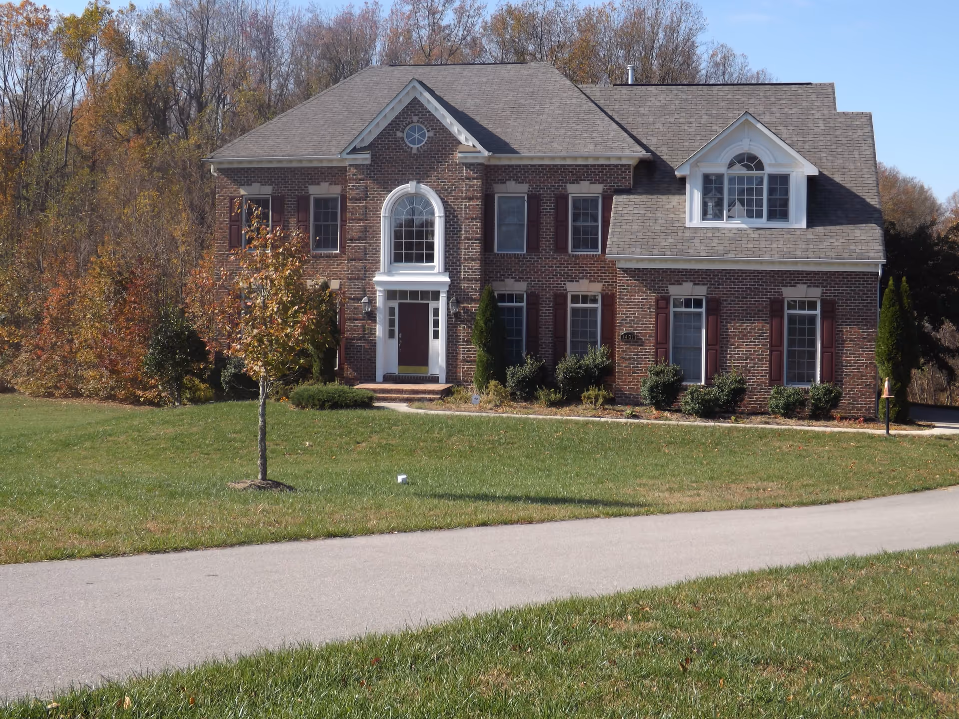 A large two-story brick house with multiple windows and a central arched window above the front door, surrounded by a green lawn and trees with autumn foliage in the background under a clear blue sky.