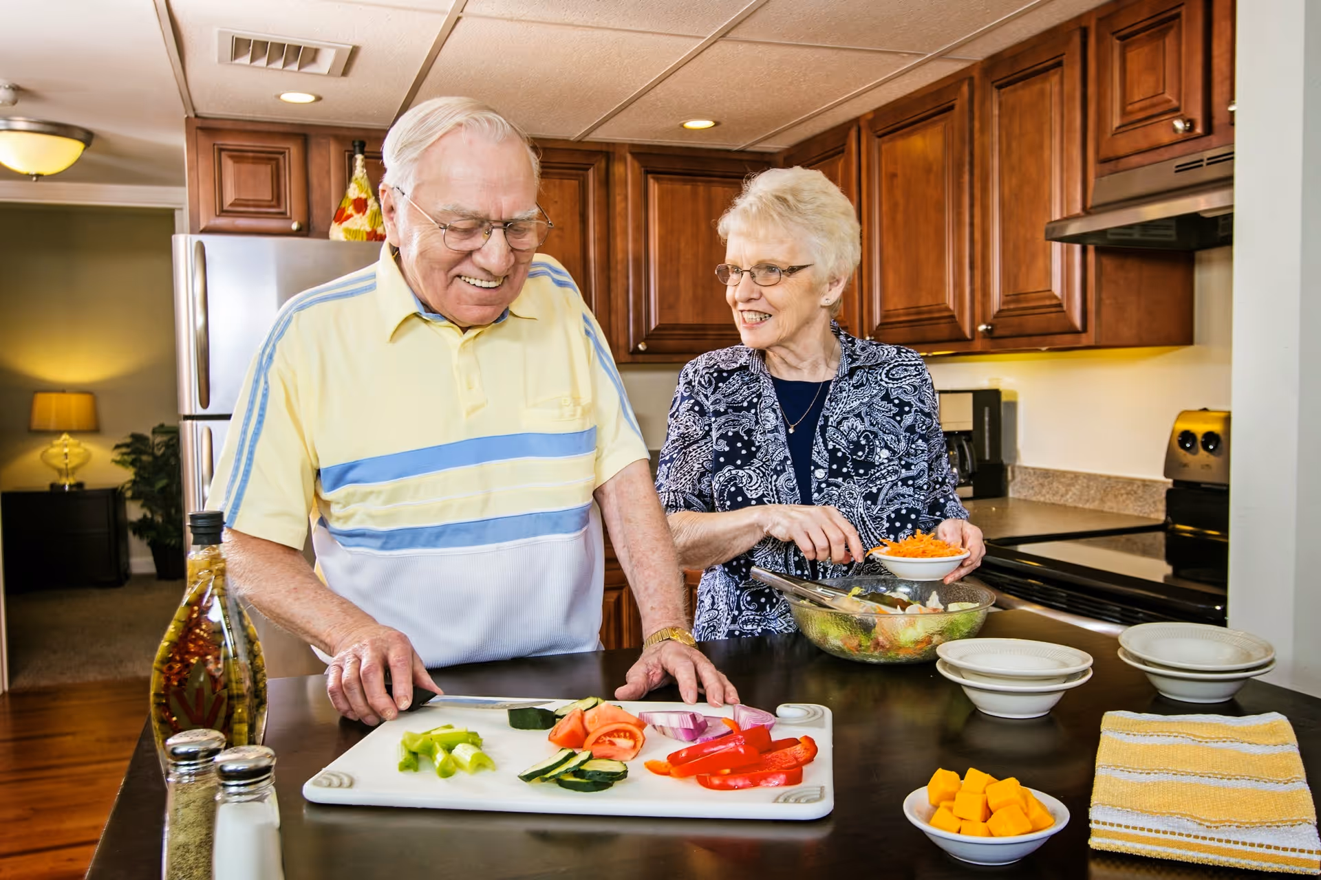 An elderly man and woman preparing a salad together in a kitchen. The man is slicing vegetables on a cutting board, while the woman is holding a bowl of shredded carrots over a large glass salad bowl. The kitchen has wooden cabinets, a stove, and a refrigerator in the background.