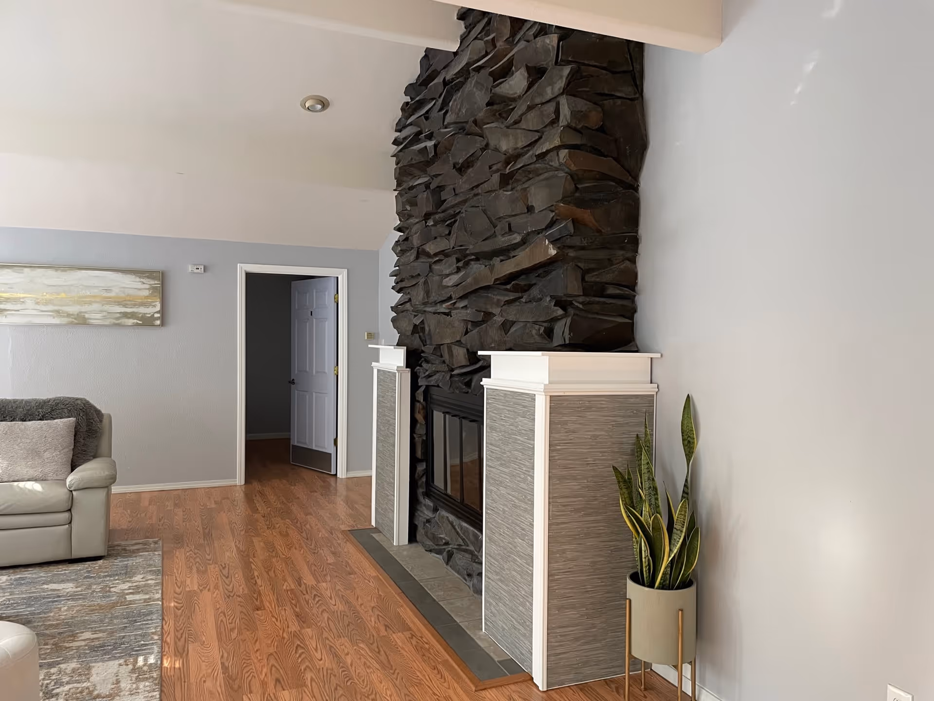 A cozy living room with a stone fireplace featuring dark, rugged stones and white mantelpieces. To the left, there is a light-colored leather sofa with a gray throw pillow and a textured rug on the wooden floor. A potted snake plant in a modern stand is placed to the right of the fireplace. The walls are painted light gray, and there is a doorway leading to another room in the background.