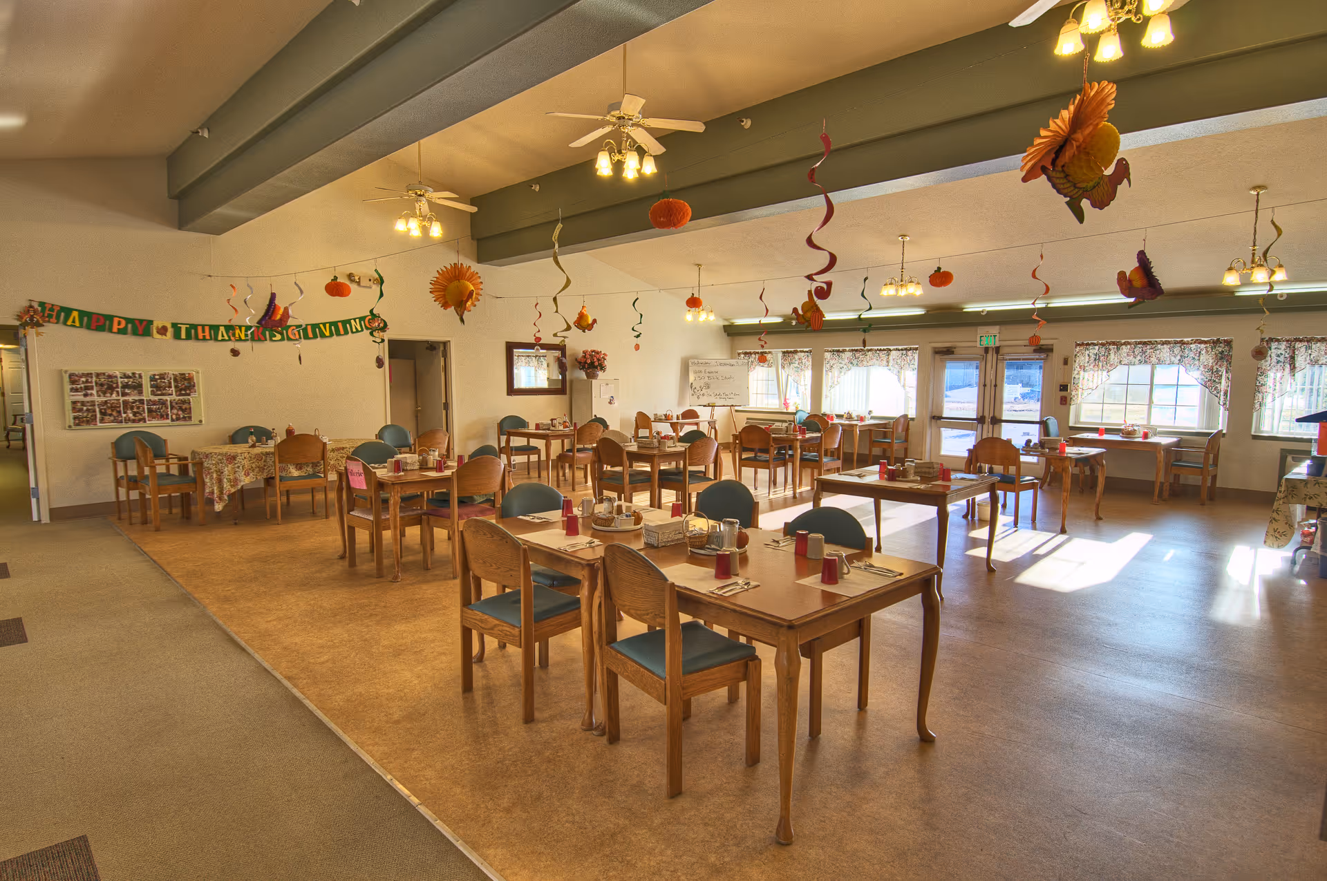 A bright dining room decorated for Thanksgiving with hanging autumn-themed decorations and a 'Happy Thanksgiving' banner. The room contains multiple wooden tables and chairs, each set with placemats, utensils, and red cups. Large windows with floral curtains allow natural light to fill the space.