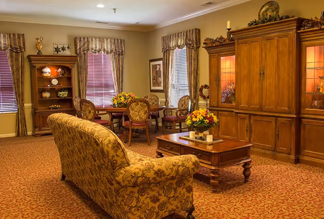 A warmly decorated living room with patterned carpet and floral upholstered furniture including a sofa and chairs around a wooden coffee table. There is a wooden cabinet with glass doors displaying decorative items, a wooden bookshelf with ornaments, and windows with striped curtains letting in natural light.