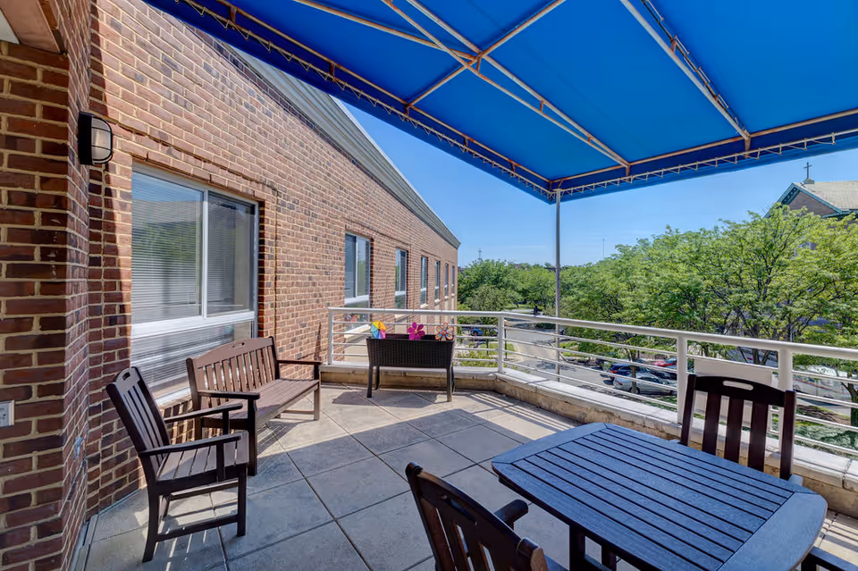 Shaded outdoor terrace with patio furniture and a blue awning overlooking trees and a parking lot beside a brick building.