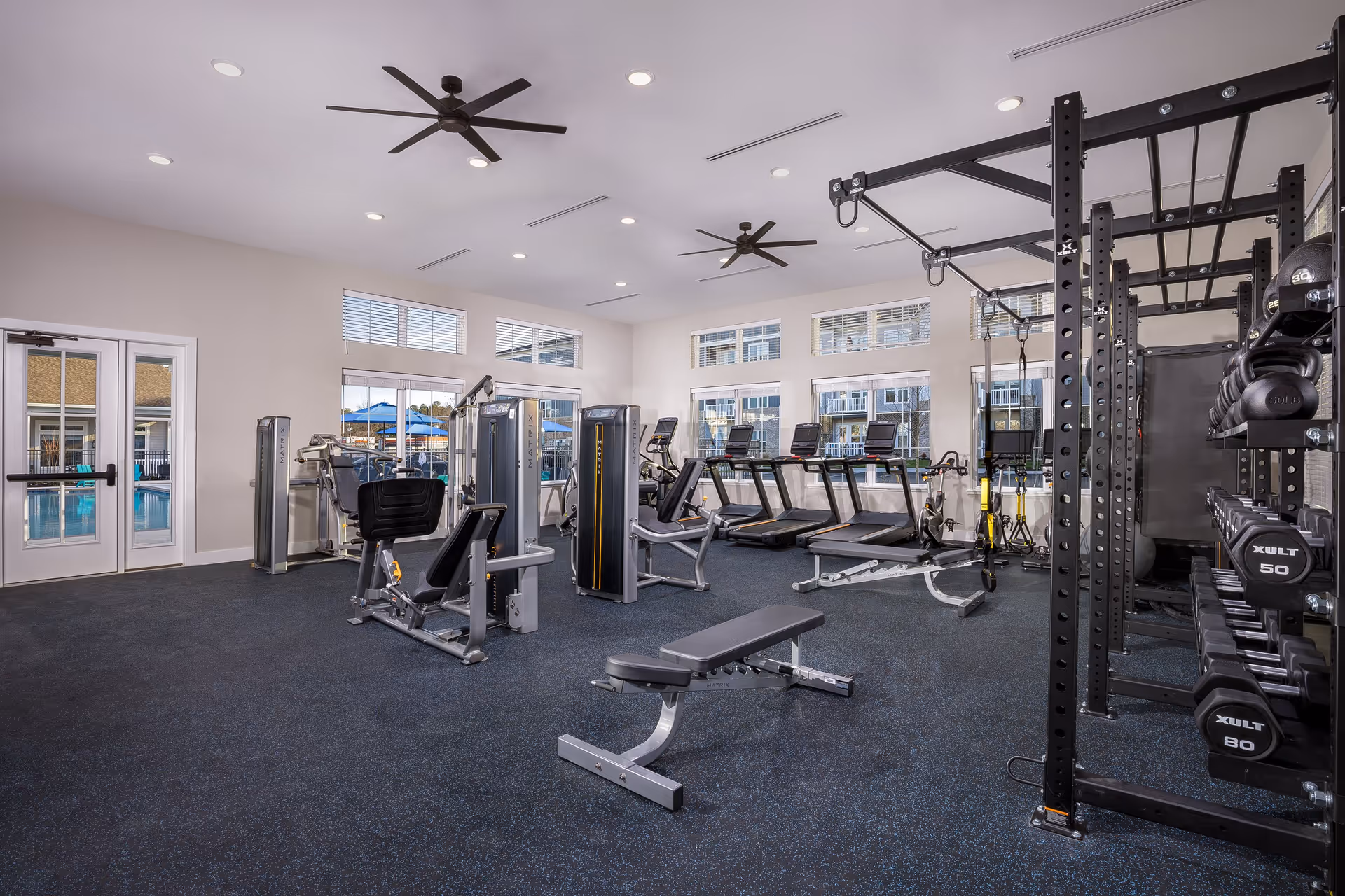 Well-lit fitness center with treadmills, weight machines, benches, and dumbbells under ceiling fans and large windows.