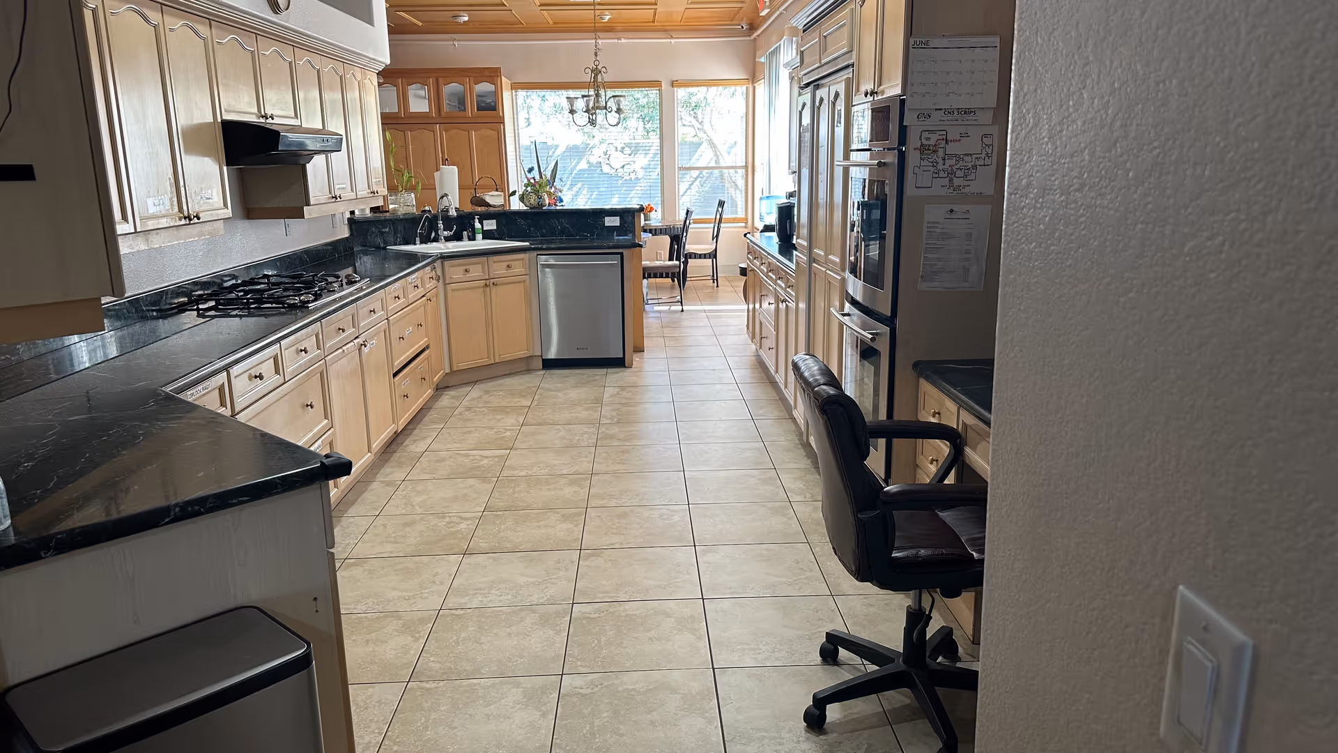 A spacious kitchen with light wood cabinets, black granite countertops, and tiled floor. The kitchen features a gas stove, dishwasher, double oven, and a desk area with a black office chair. At the far end, there is a dining area with a table and chairs near large windows letting in natural light.