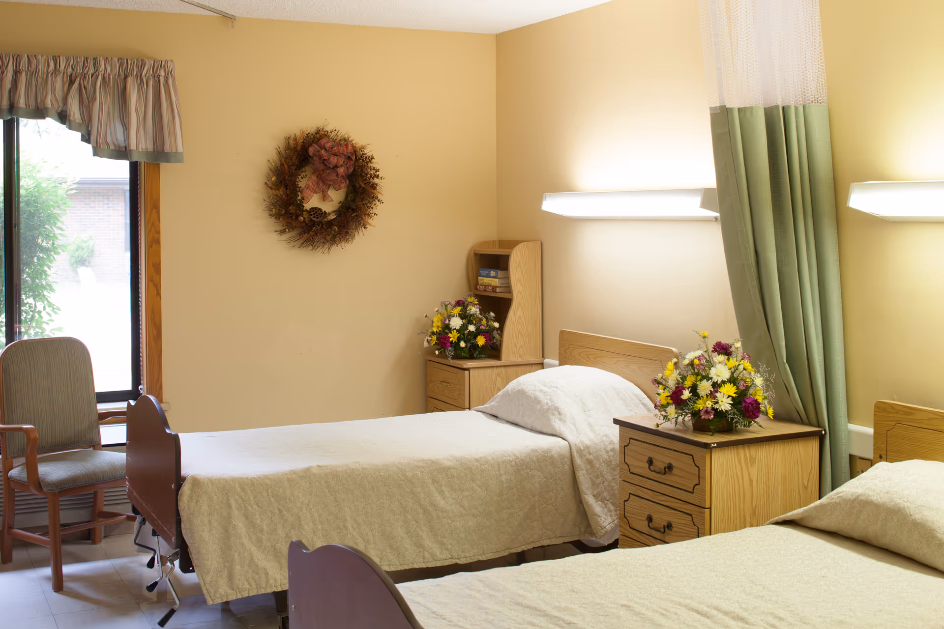 A room with two single beds separated by a wooden nightstand with a flower arrangement. Each bed has a beige bedspread and a pillow. There is a green privacy curtain partially drawn between the beds. On the wall above each bed is a long horizontal light fixture. A wooden chair is positioned near a window with a valance curtain. A wreath hangs on the beige wall above a small wooden cabinet with another flower arrangement and some books.