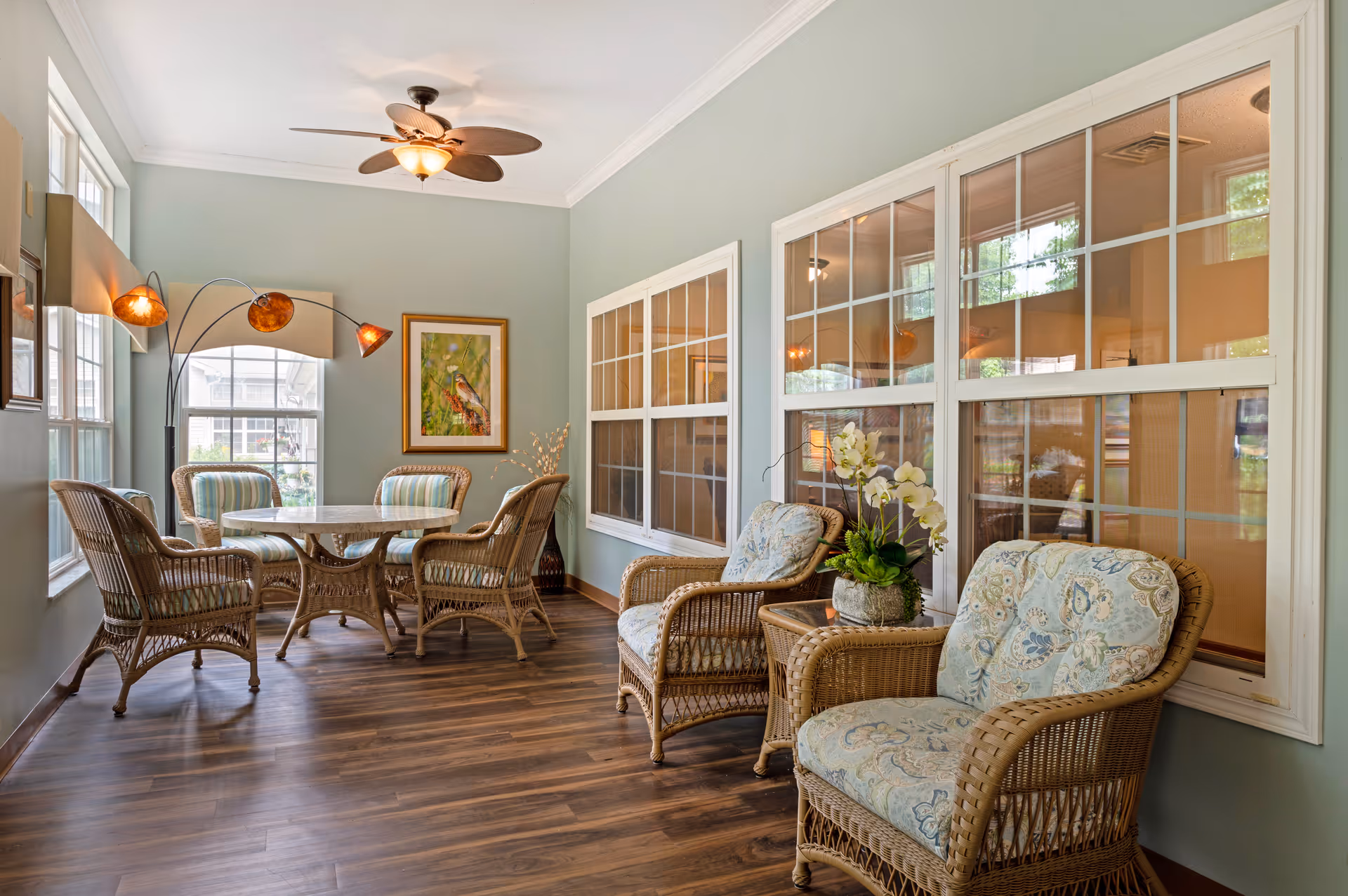 Sunlit sitting area with wicker armchairs and a round table beside large windows.