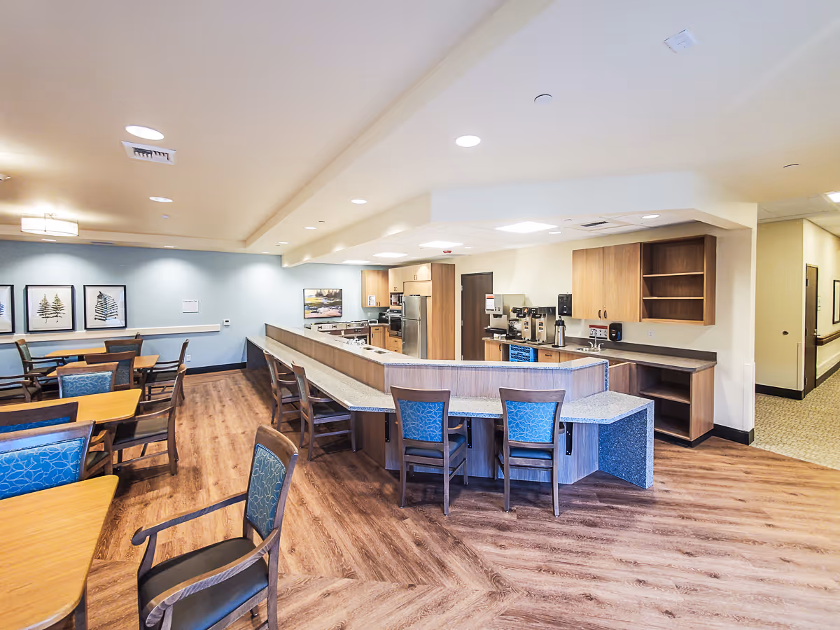 A spacious dining area with wooden tables and chairs featuring blue patterned cushions. The room has a light blue accent wall with framed artwork and a kitchen area with wooden cabinets, a refrigerator, coffee machines, and a long counter with additional seating. The floor is wood-patterned, and the ceiling has recessed lighting.