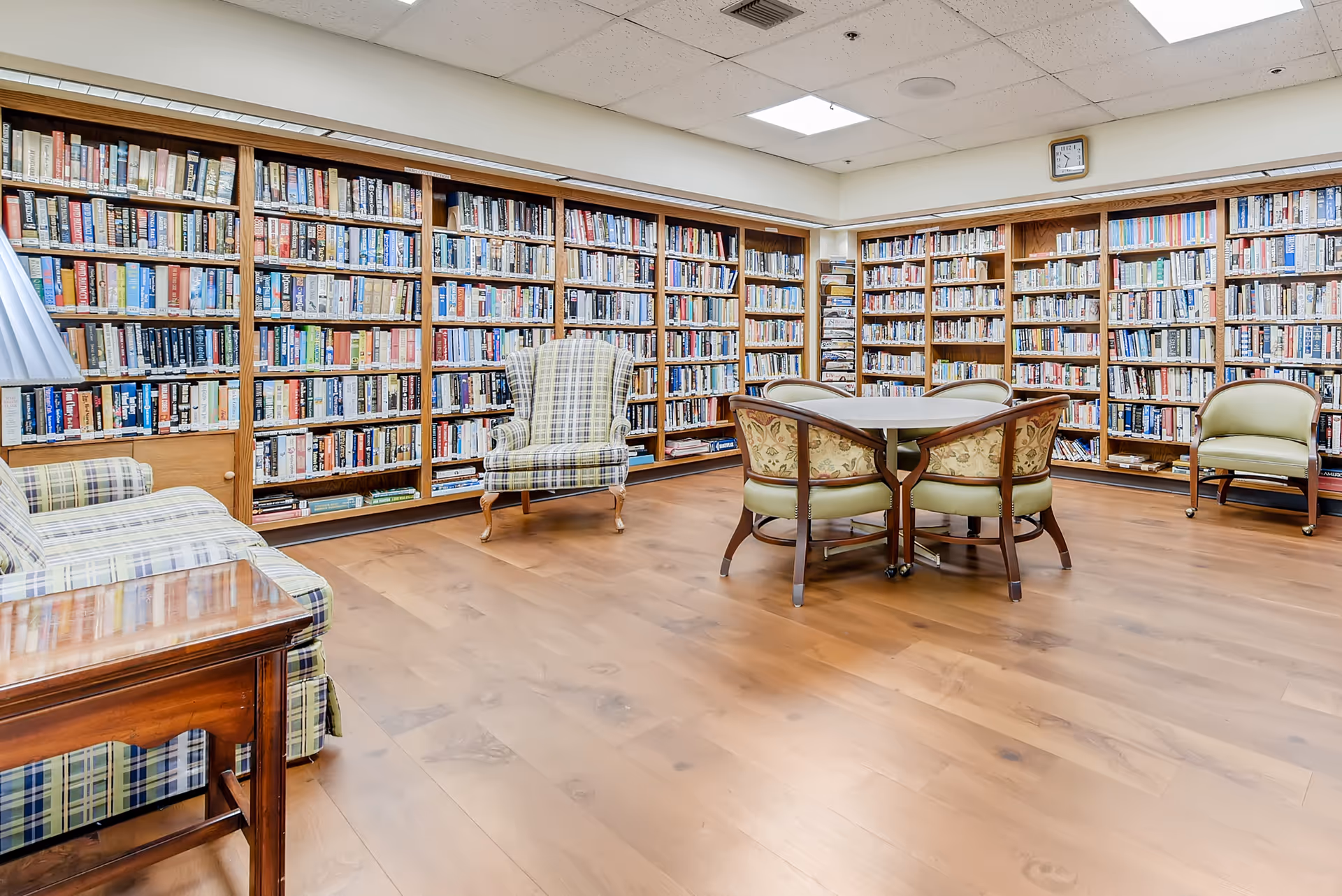 A senior living facility library room with wooden bookshelves filled with books along the walls, a wooden floor, a table with four chairs in the center, and several upholstered armchairs and a sofa with plaid fabric.