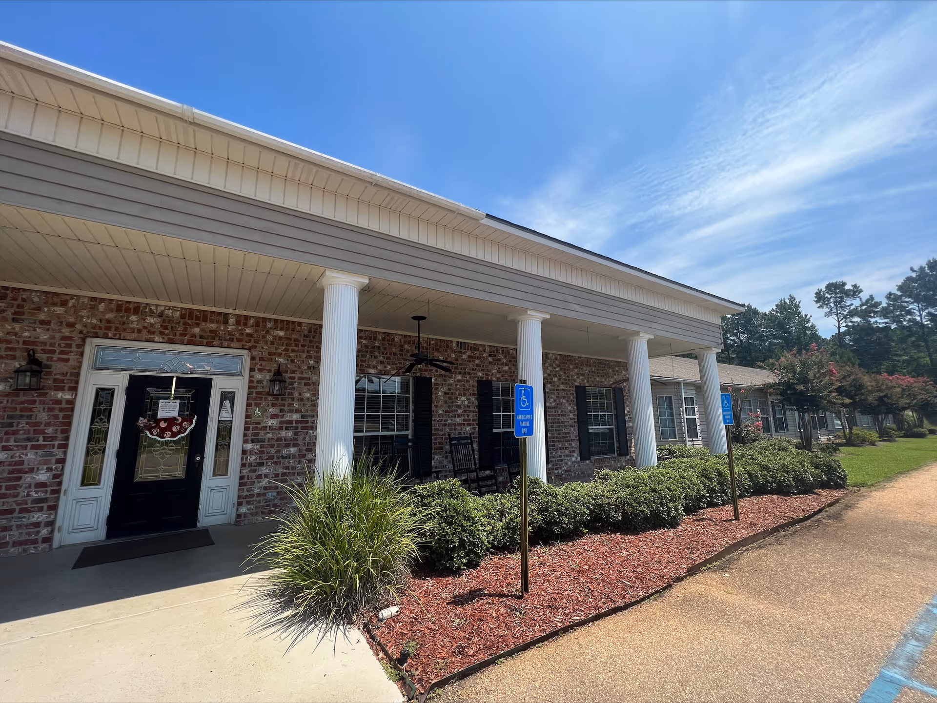 Exterior view of a single-story brick building with white columns supporting a covered porch. The entrance features a black door with decorative glass panels and a wreath. There are two handicap parking signs in front of the building, along with neatly trimmed bushes and a mulched garden bed. The sky is clear with some wispy clouds.